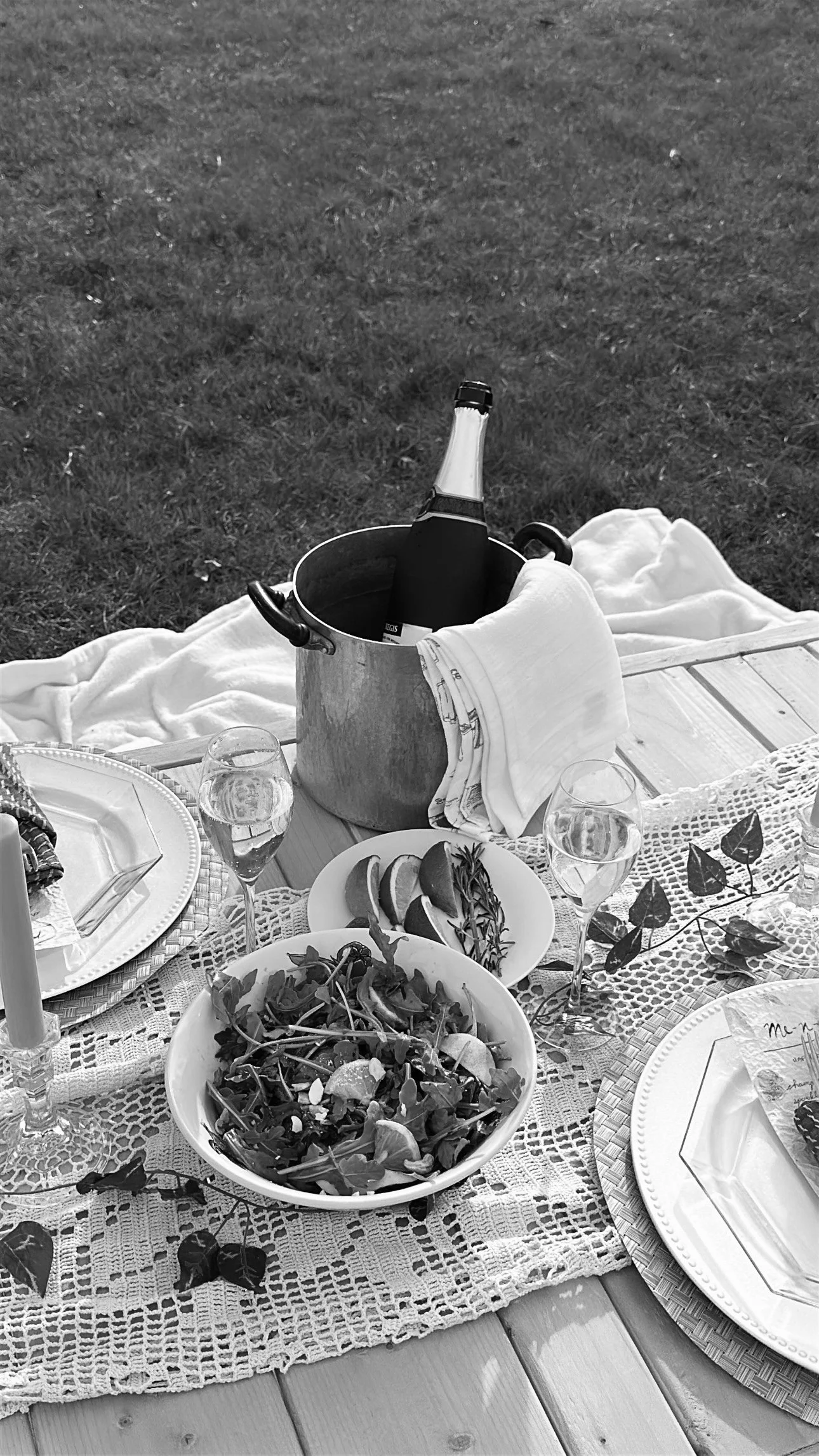 A table set for an outdoor meal with a bowl of salad, a plate with slices of lemon, wine glasses, and a bottle of champagne in an ice bucket, on a lace table runner.