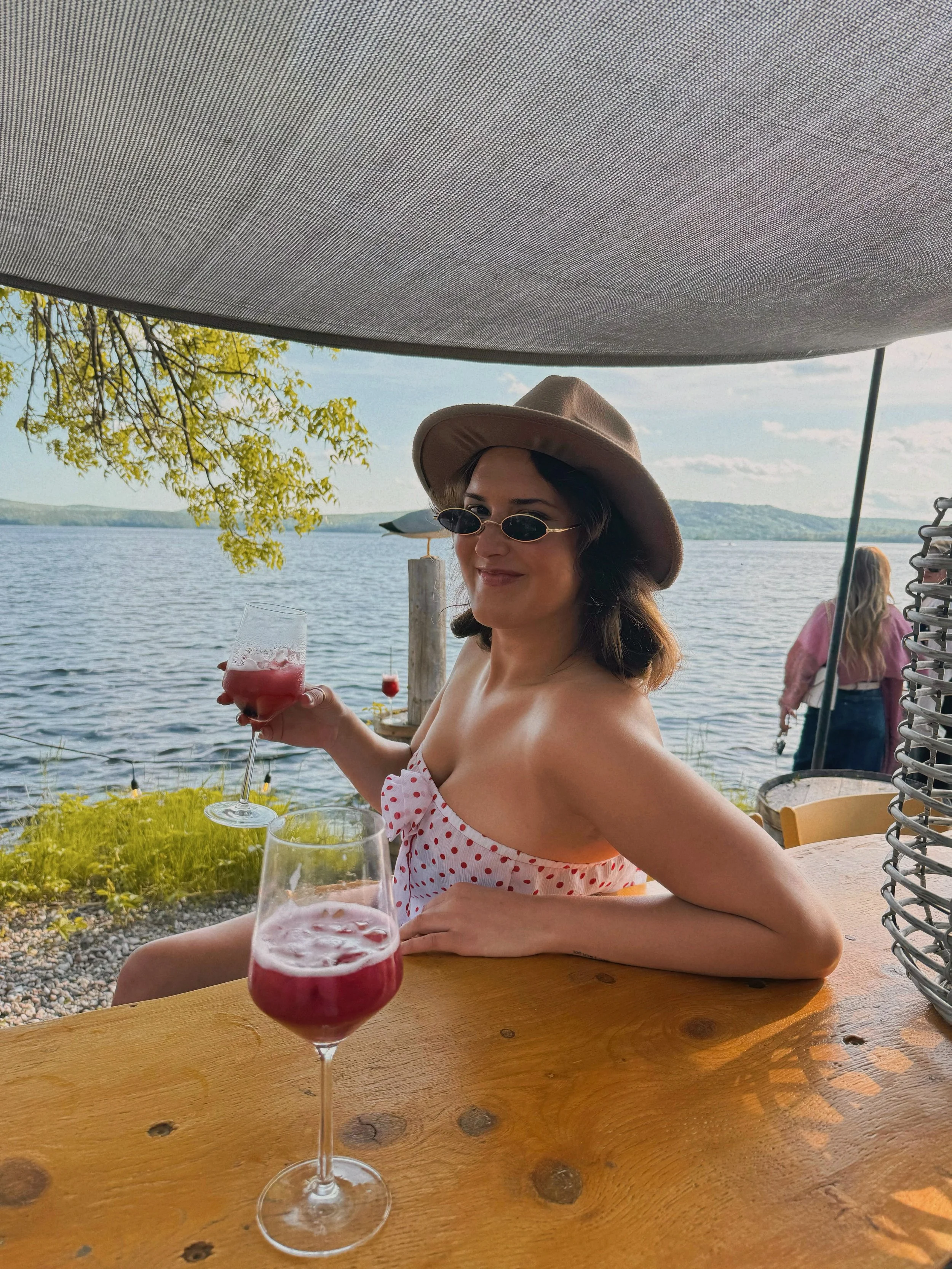 A woman sitting at a table near a lake, holding a glass of red wine or cocktail, wearing sunglasses, a wide-brimmed hat, and a polka dot strapless top, with an outdoor setting and another person in the background.