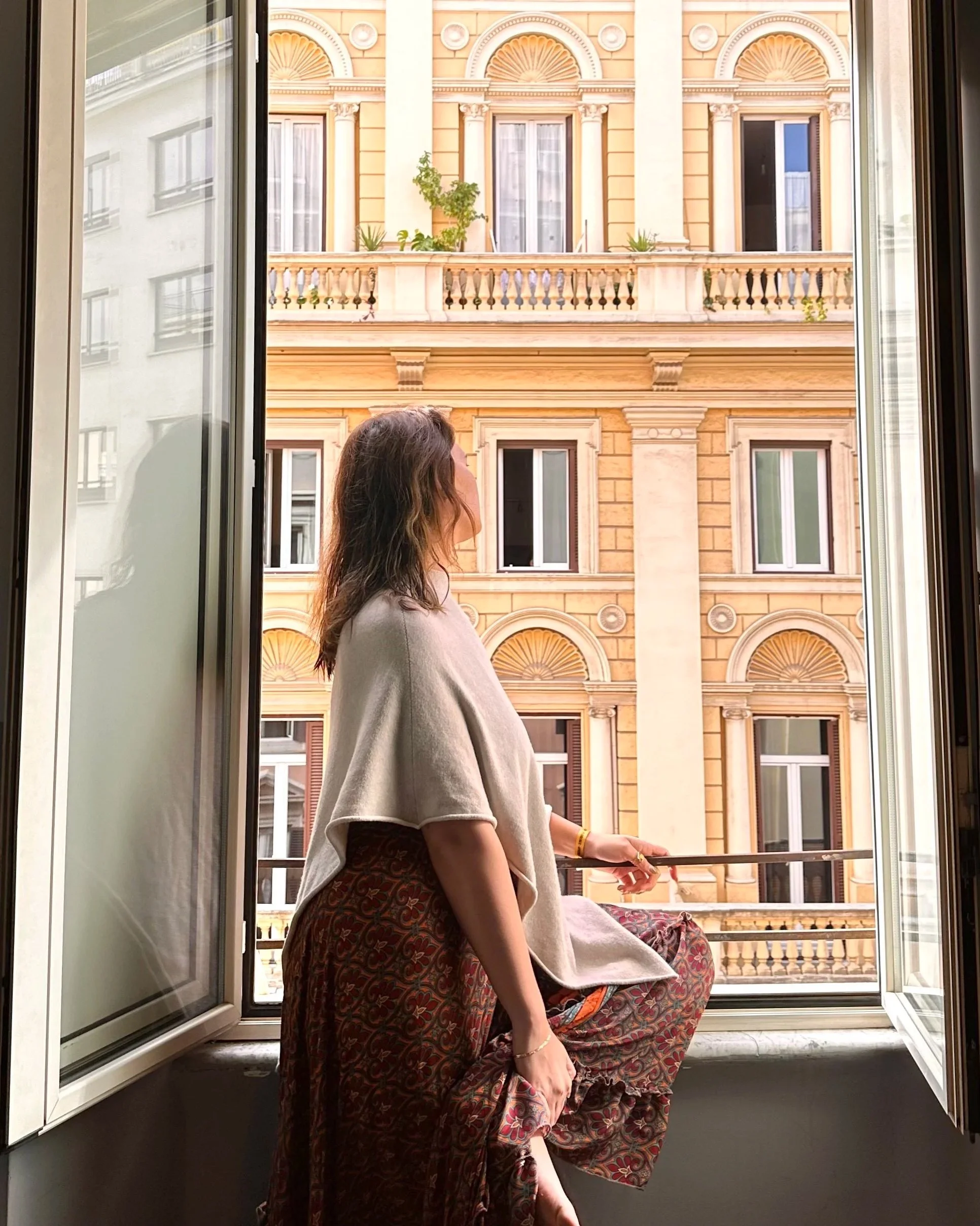 Woman sitting on windowsill looking outside at a building with ornate architecture and large windows, with some plants on the balcony.