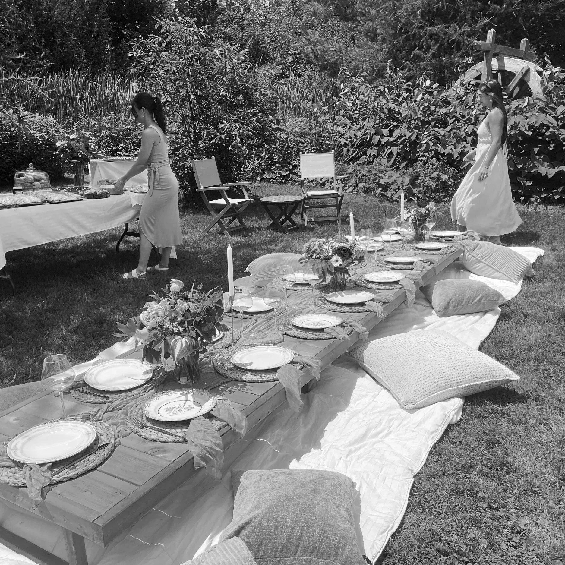 Outdoor garden party setup with a low wooden table decorated with flowers, candles, and dinnerware on pillows and blankets on the grass. Two women in dresses are arranging food and decorations amid trees and greenery.