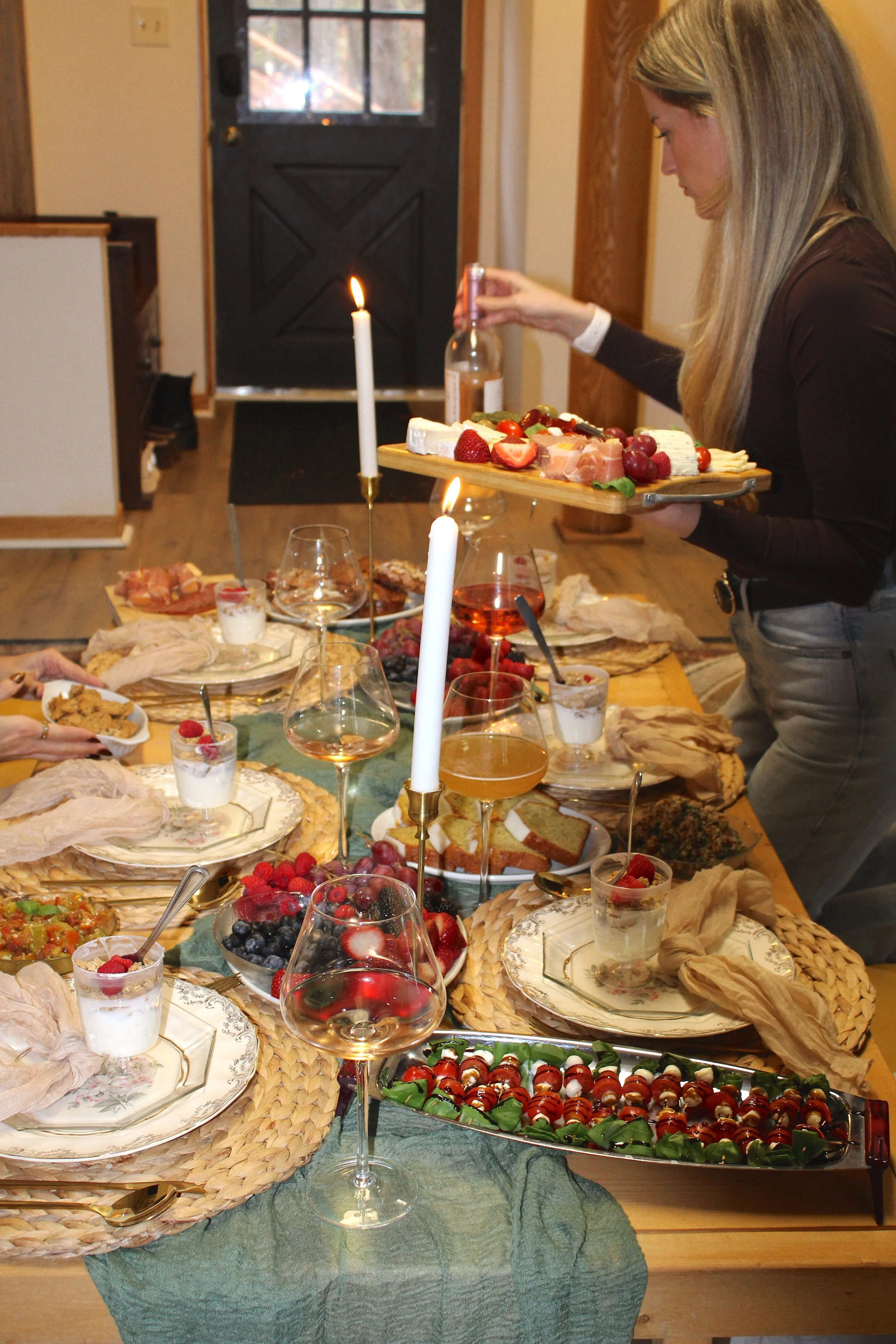 A woman preparing a charcuterie and dessert board at a dining table with wine glasses, candles, and various fruits and snacks.