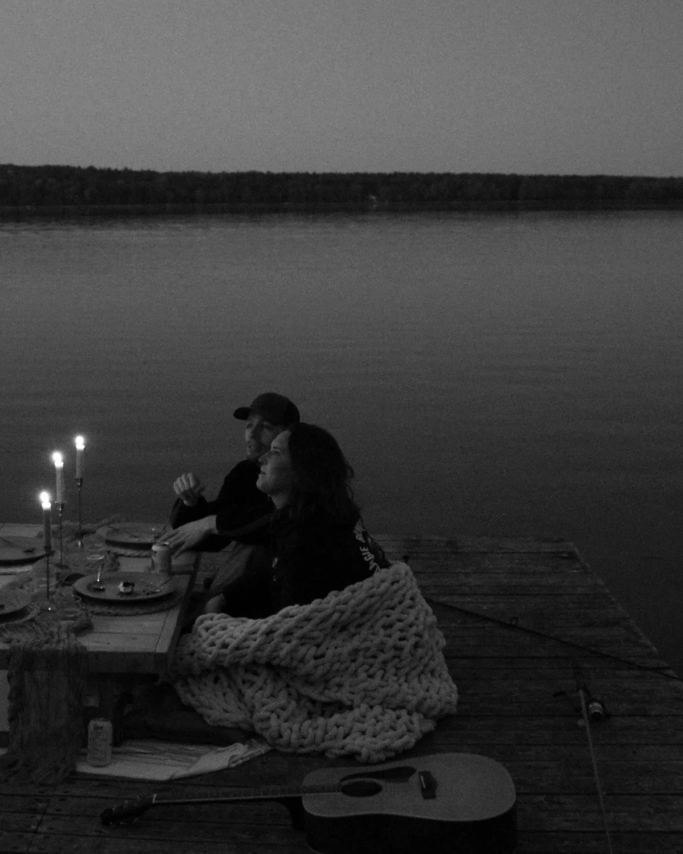 A couple sitting at a table on a dock by a lake during evening with candles and food, one person is smiling and wearing a baseball cap, the other is wrapped in a large chunky knit blanket.
