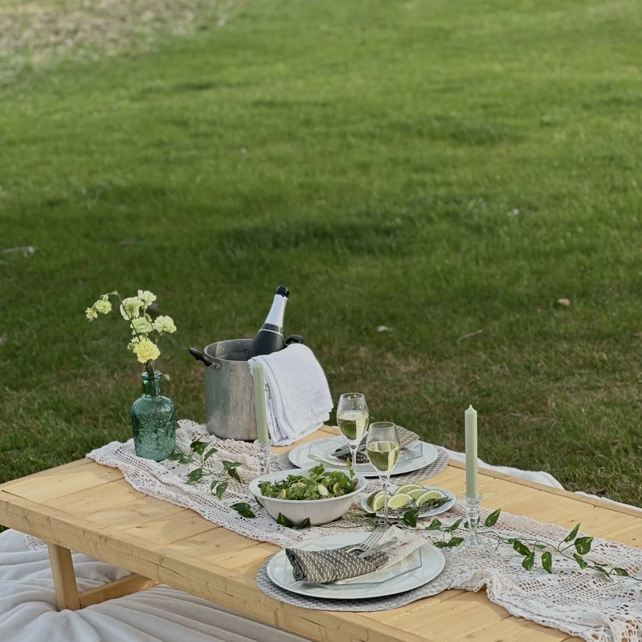 A picnic setup on a wooden table outdoors, featuring a flower vase with yellow and white flowers, a champagne bucket with a bottle, two champagne glasses, a salad bowl, a plate with lime slices, a candlestick with a white candle, and plate settings with napkins and silverware, on a white blanket with green grass in the background.