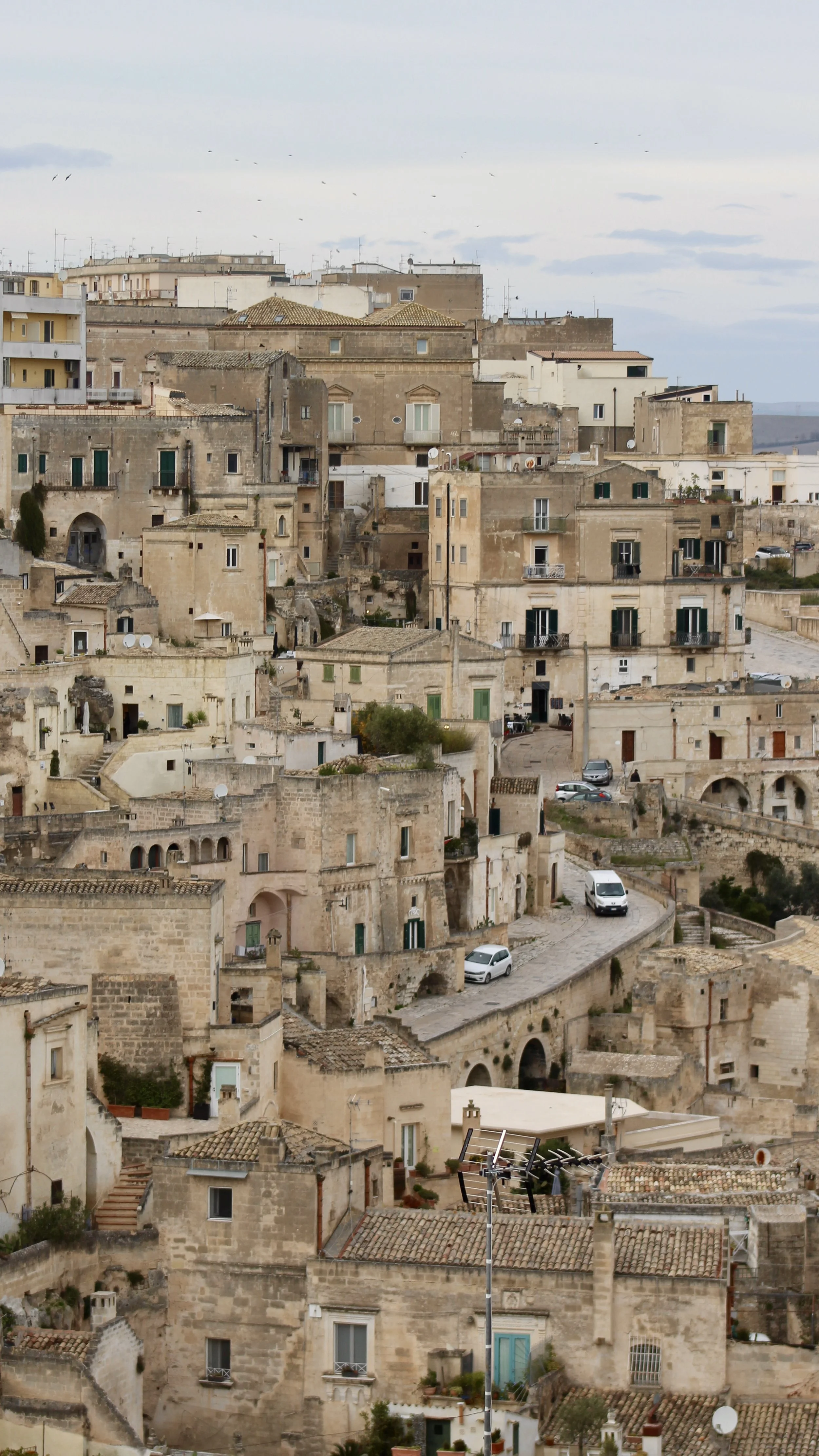 A hillside view of beige stone buildings with narrow winding roads, some cars, and satellite dishes, in a historic Mediterranean town.