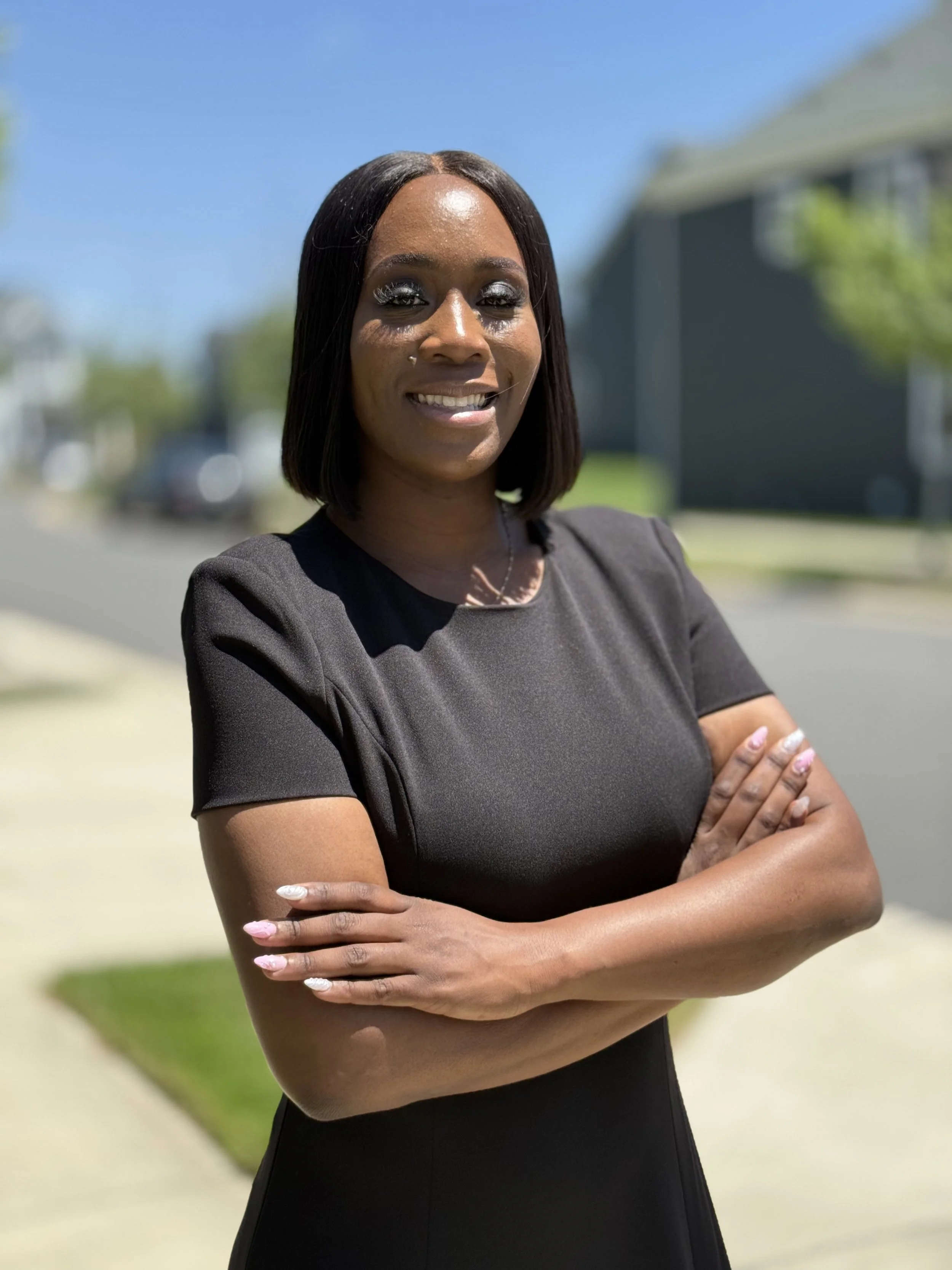 A woman with shoulder-length black hair stands outside on a sunny day with her arms crossed, smiling at the camera. She is wearing a black dress and has painted nails.