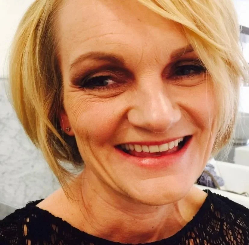 Close-up of a woman with blonde hair, smiling with visible teeth, wearing makeup, earrings, and a black lace top.
