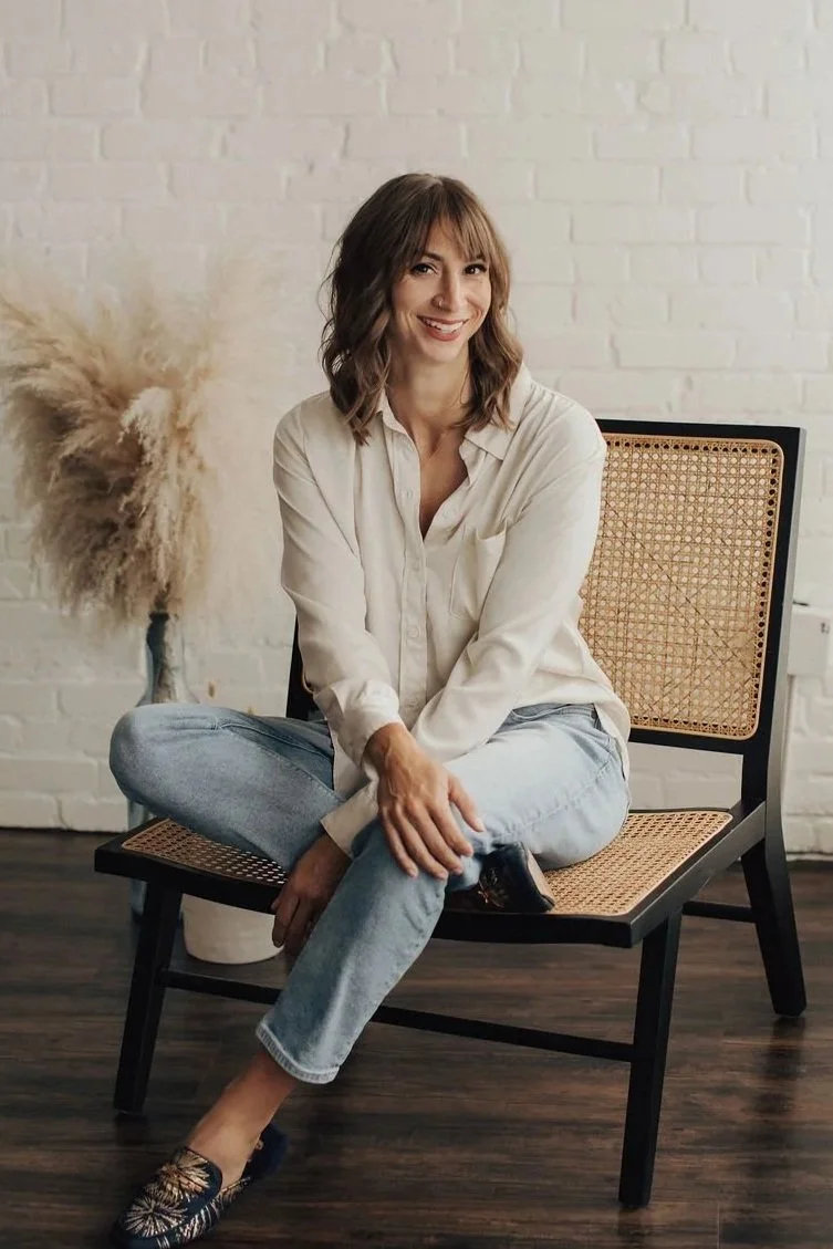 Smiling woman sitting cross-legged on a black chair with cane seat, wearing a beige shirt, light jeans, and patterned shoes, in front of a white brick wall and a vase with pampas grass. Alena Brown, the divorce coach