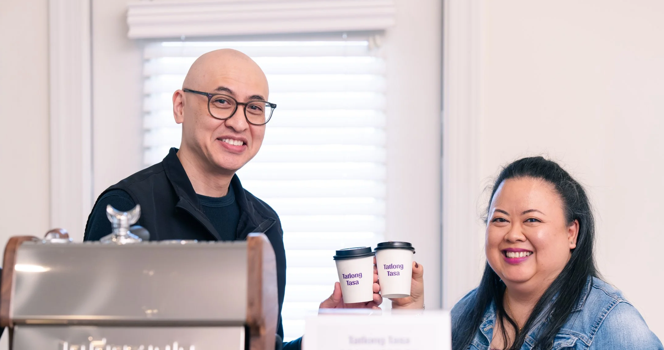 A man and woman baristas holding custom branded cups with ‘Tatlong Tasa’ behind a mobile espresso bar at a coffee cart catering event.