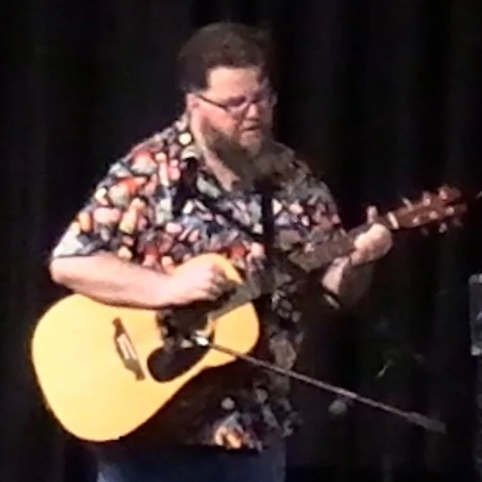 A bearded man with brown hair and glasses wearing a Hawaiian shirt and playing guitar.