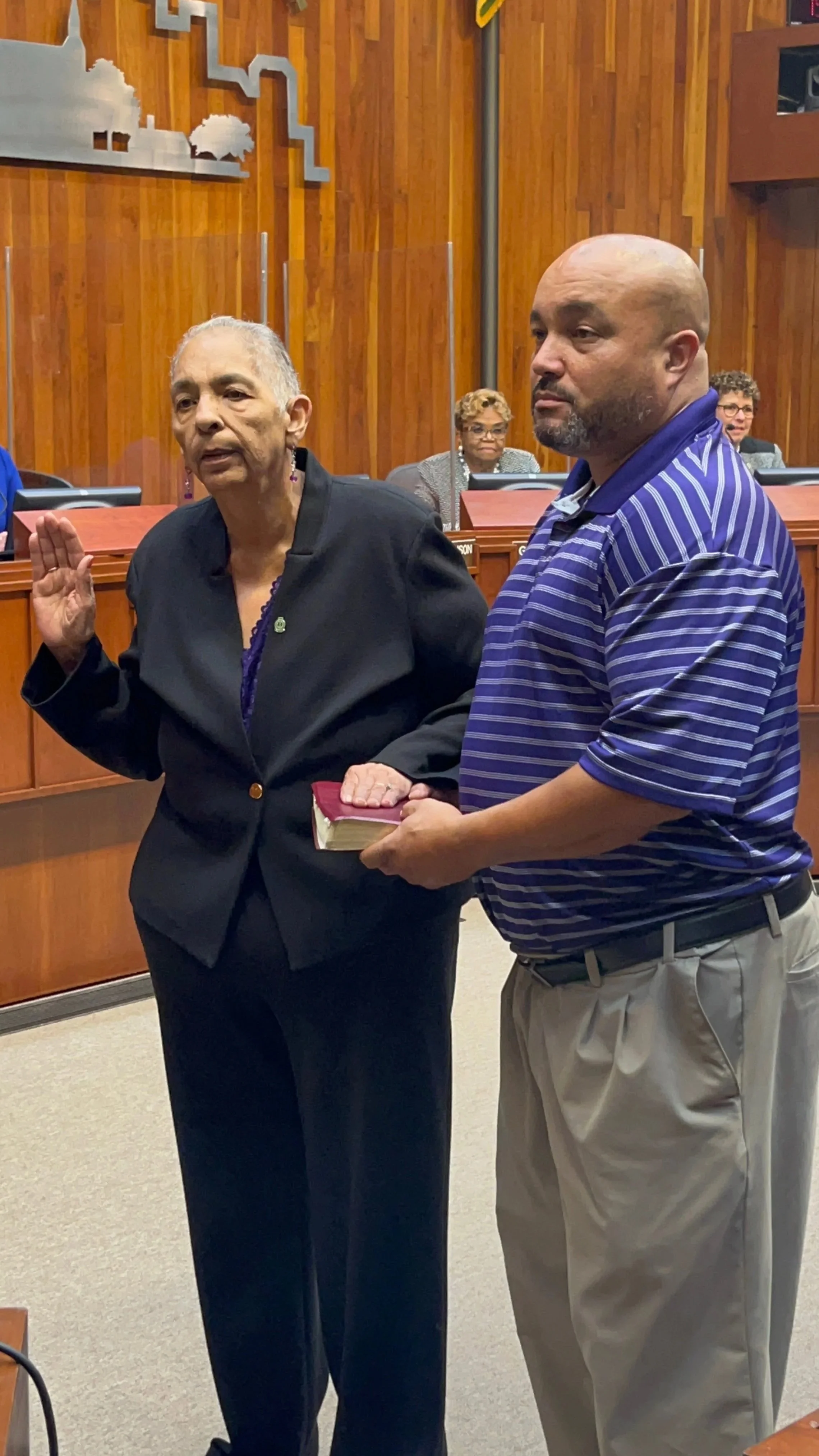 An older woman wearing a black blazer with earrings raising her right hand while holding a book in her left hand, standing next to a man in a blue striped polo shirt holding her hand. They are in a formal setting with wood-paneled walls and other women seated at a long desk in the background.