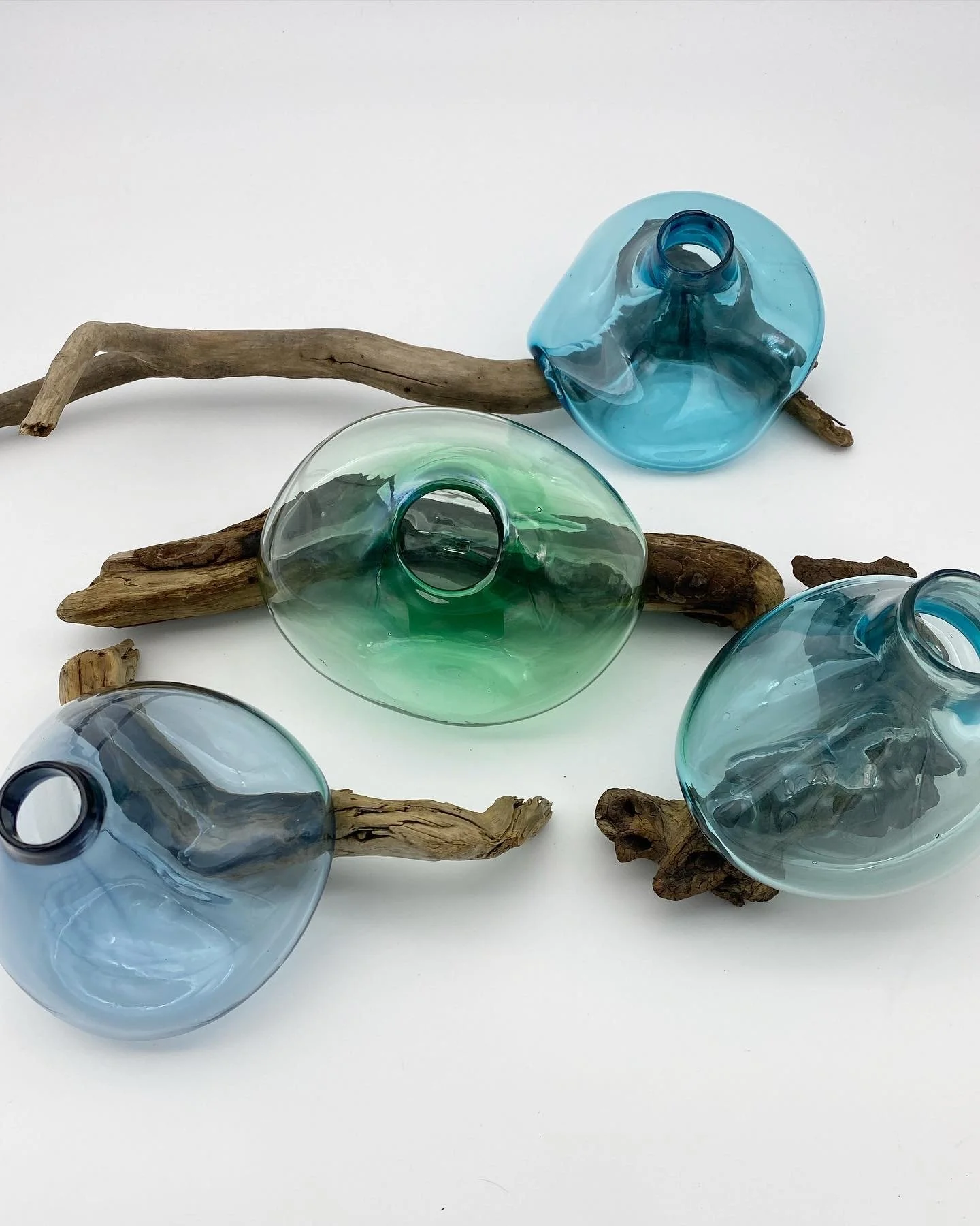 Multiple colored blown  glass vases in blue and green shades arranged on and around weathered driftwood pieces on a white background.