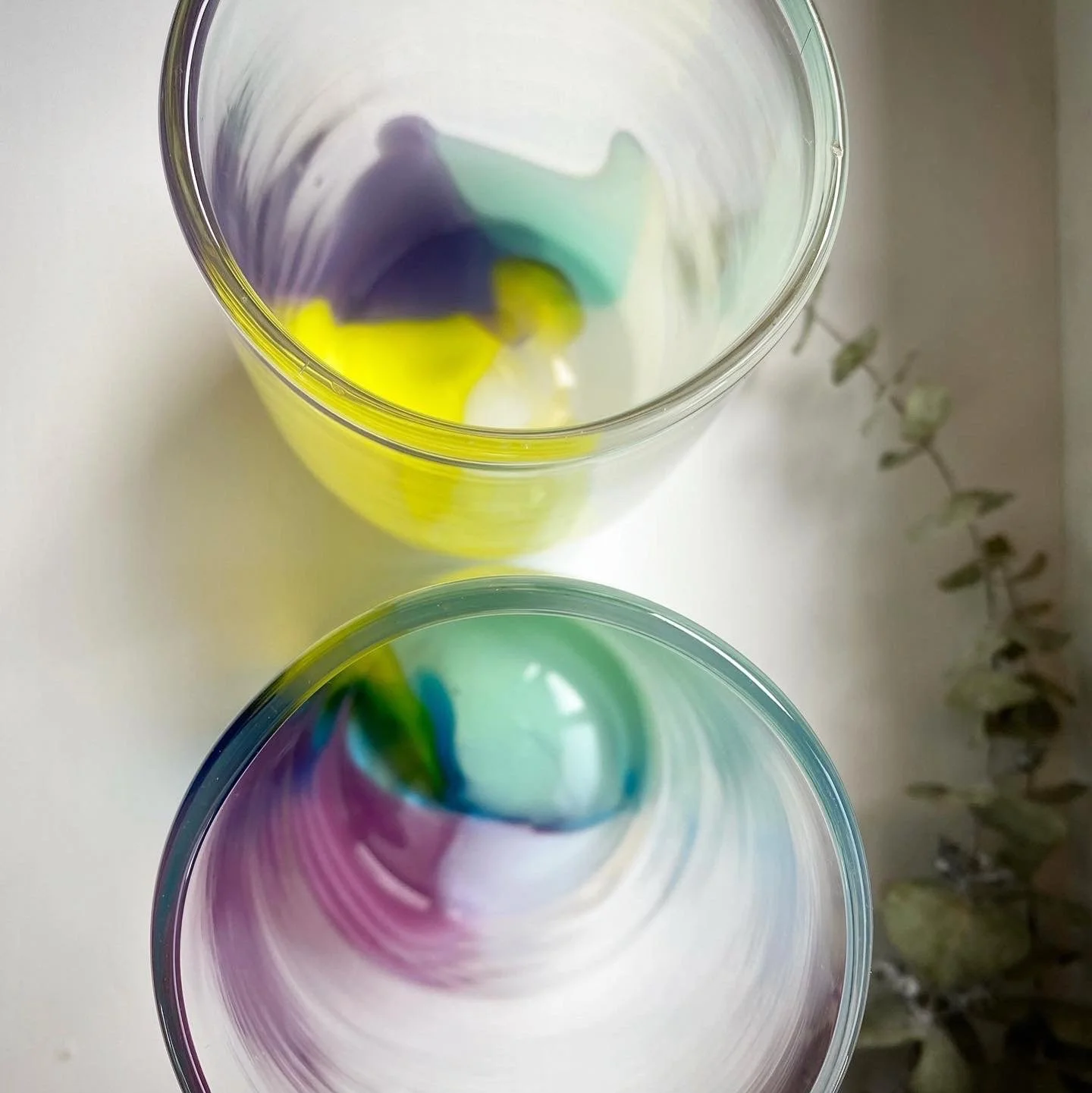 Two colorful Colour POP cups glasses with swirling abstract rainbow-like patterns inside, placed on a white surface with some greenery to the side.