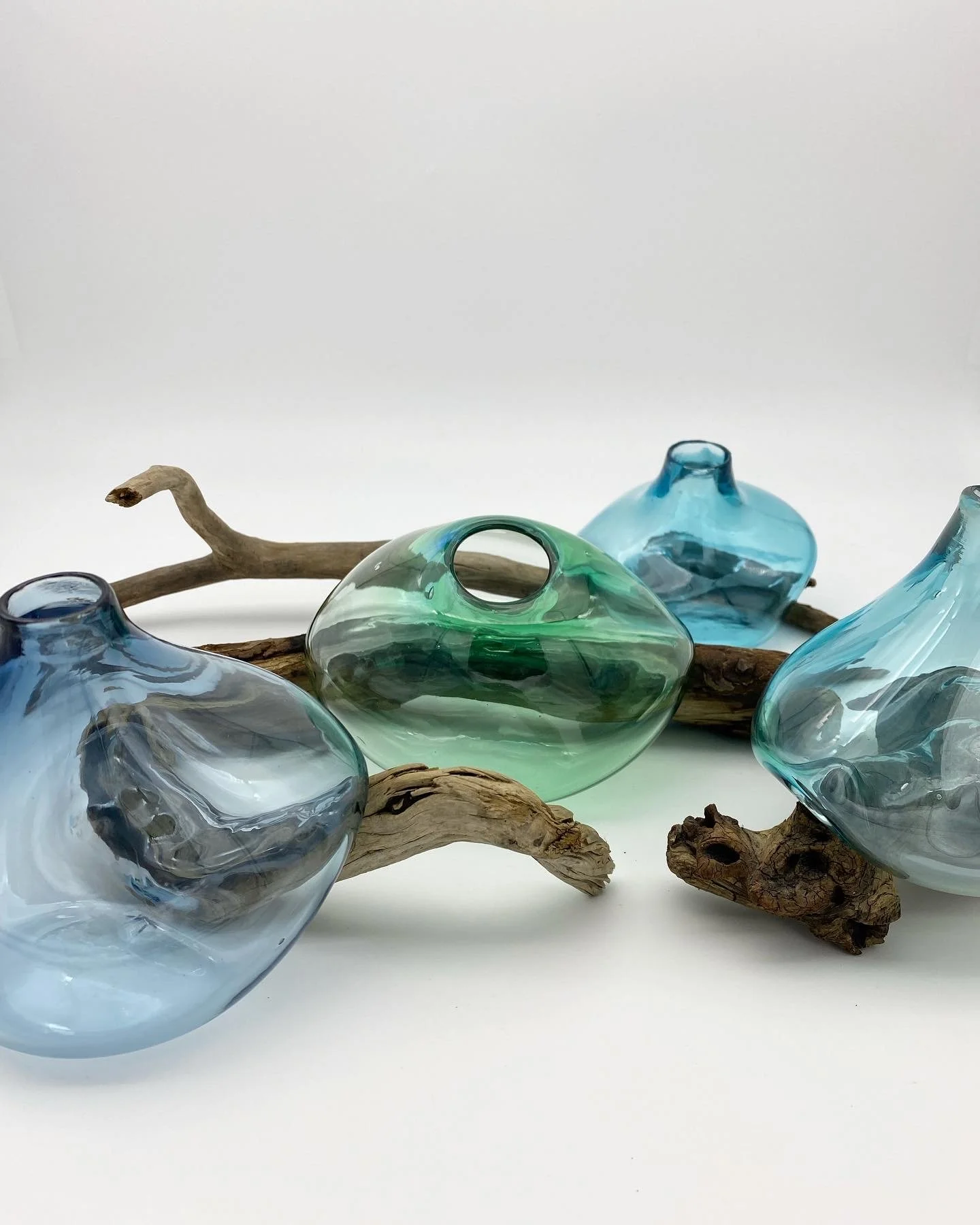 A collection of blown glass vases in shades of blue and green, resting on driftwood branches against a plain white background.
