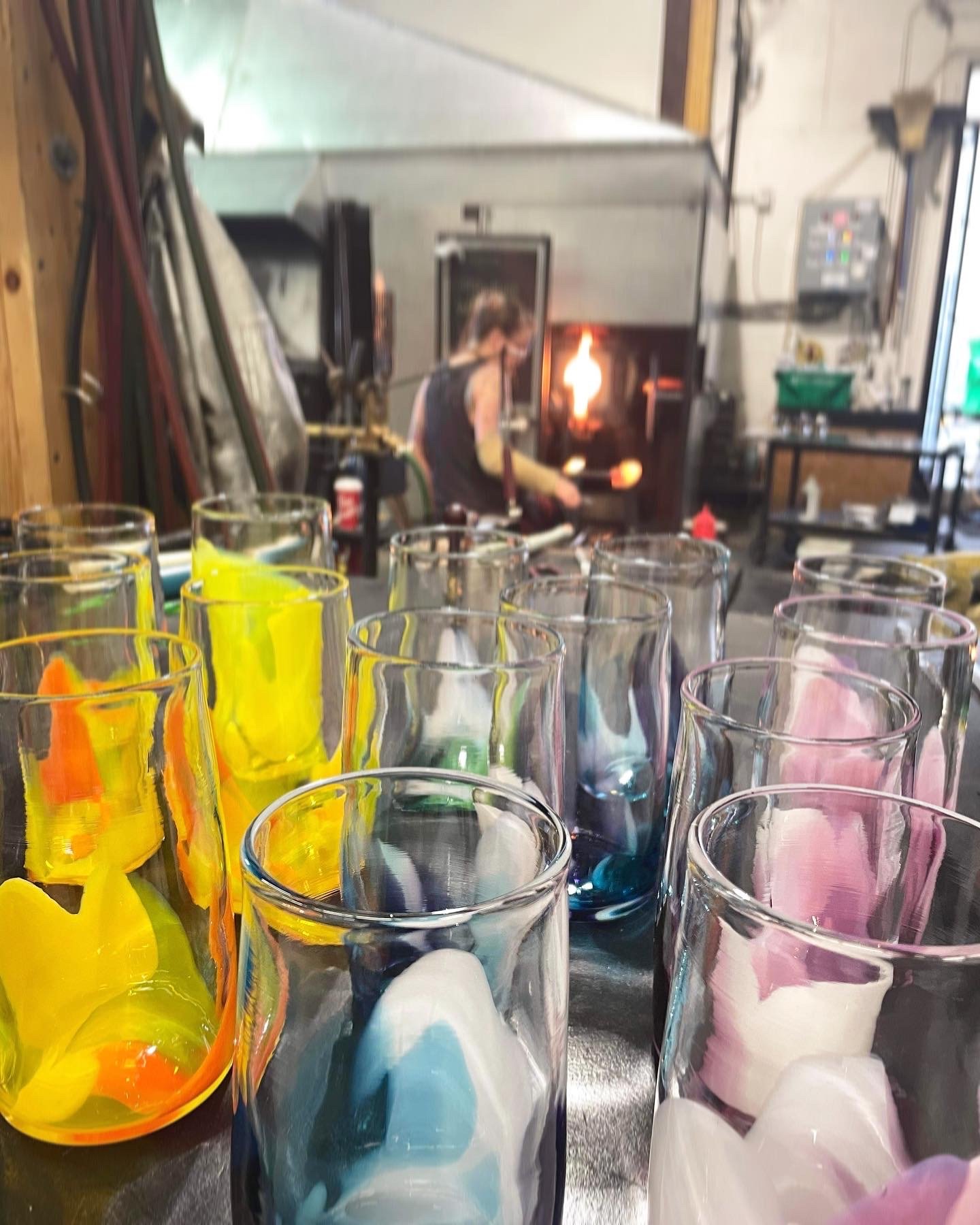 A glassblowing studio with colorful colour pop cups glass pieces in the foreground and tara working with a flame in the background.