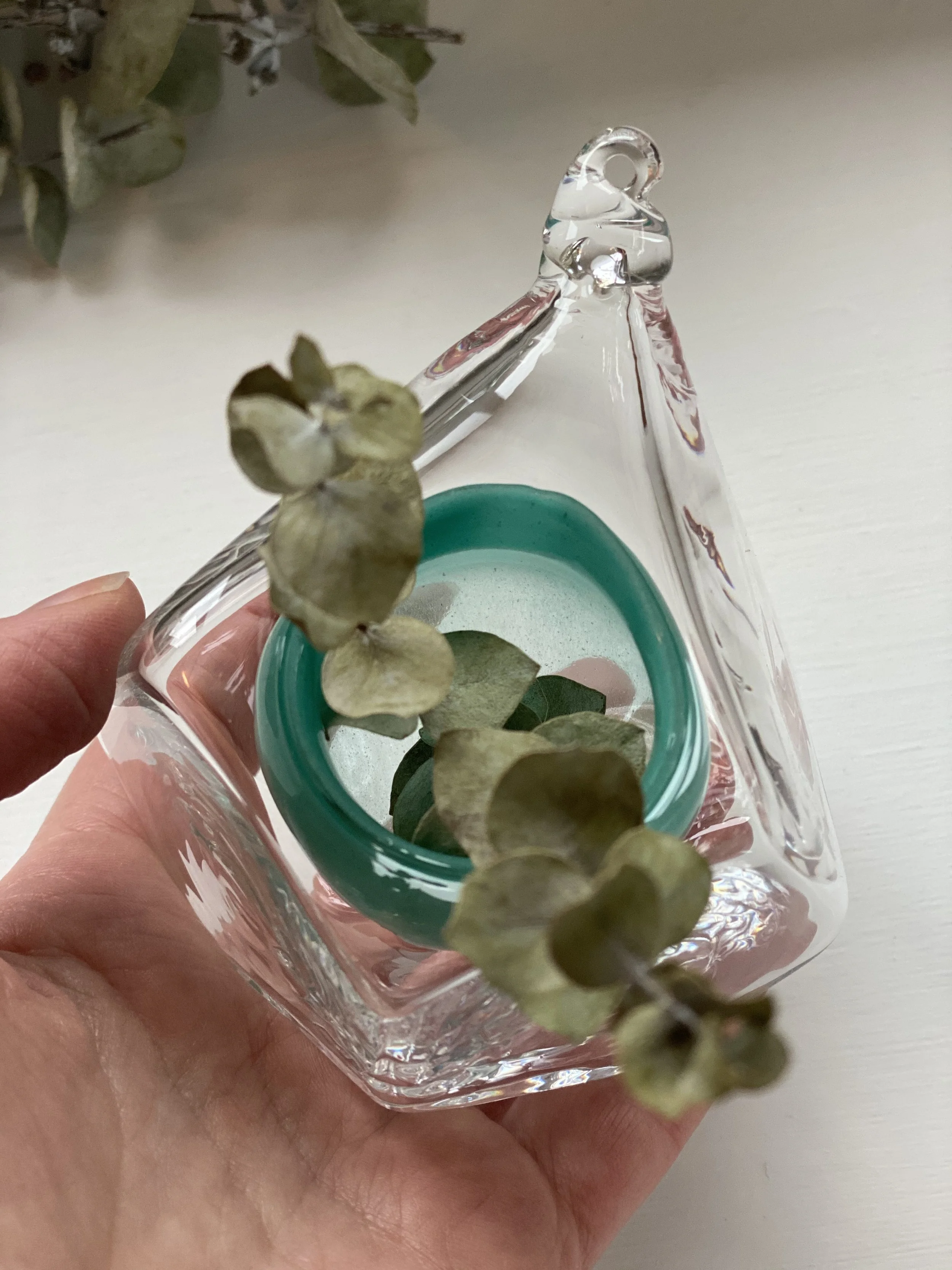 Close-up of a hand holding a small glass container with dried eucalyptus leaves inside, on a white surface.