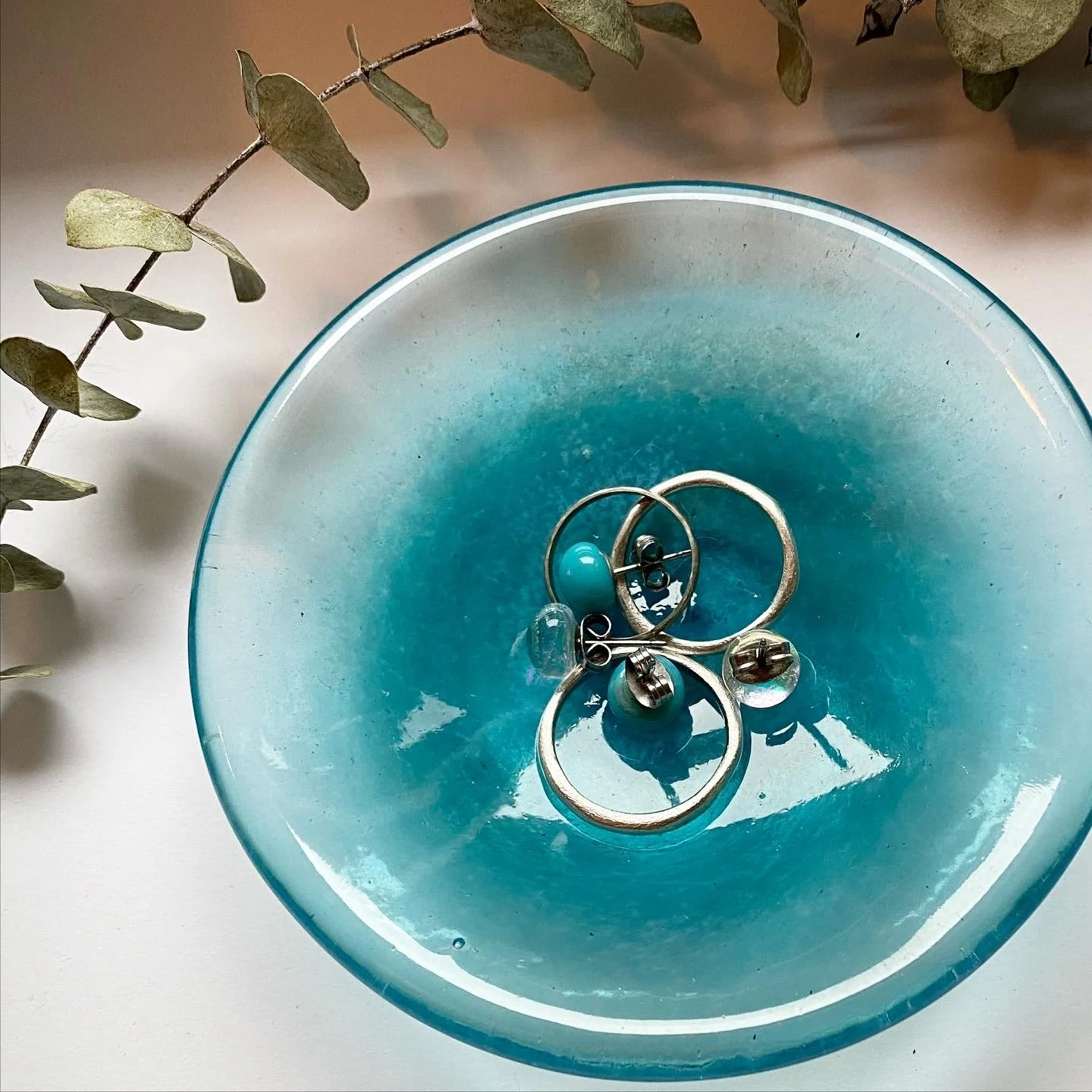 A light blue glass bowl holding various jewelry pieces, including rings and earrings, with a sprig of eucalyptus leaves nearby.