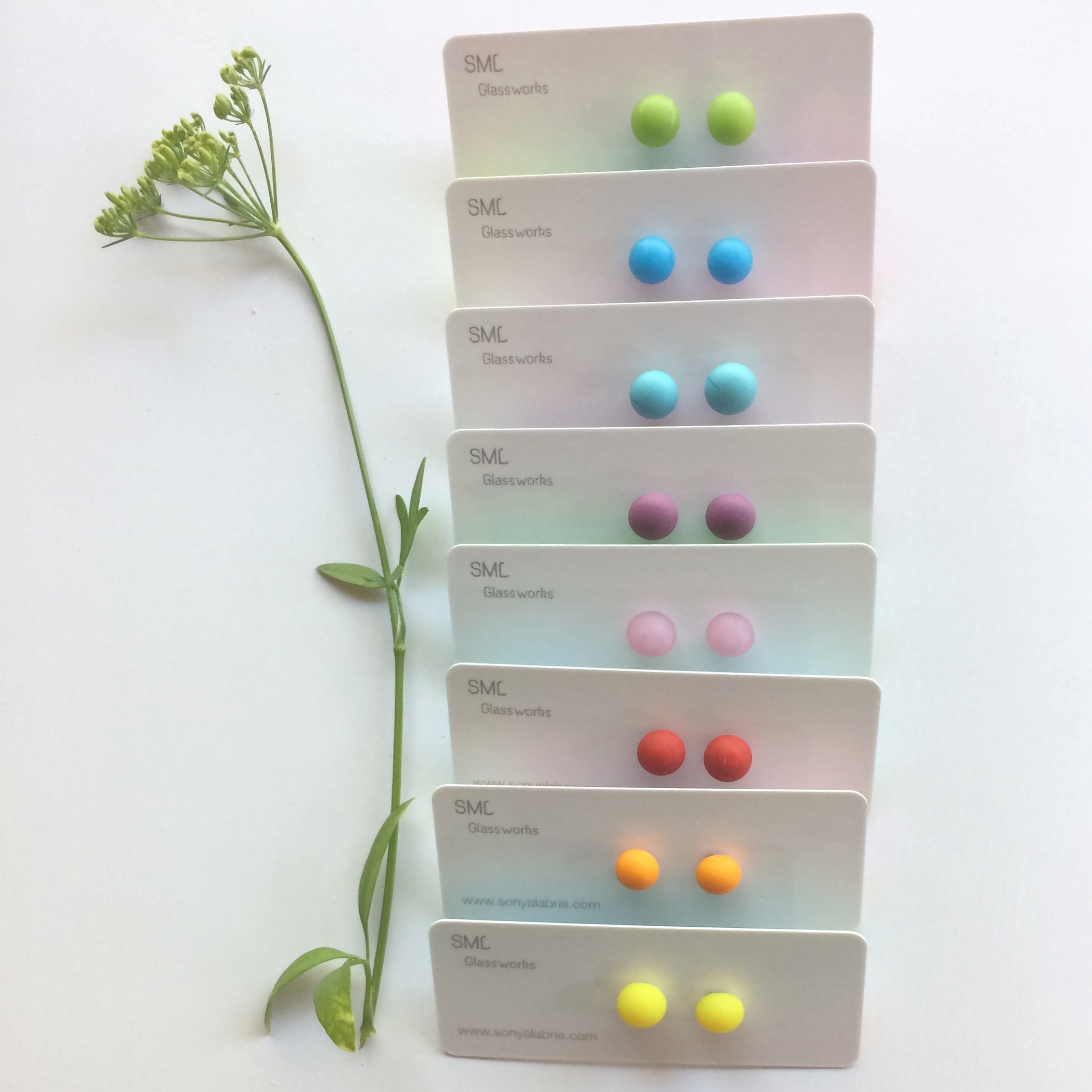 A sprig of greenery with small flowers next to three cards each displaying a pair of colorful round earrings in green, blue, pink, and yellow, arranged vertically on a white background.