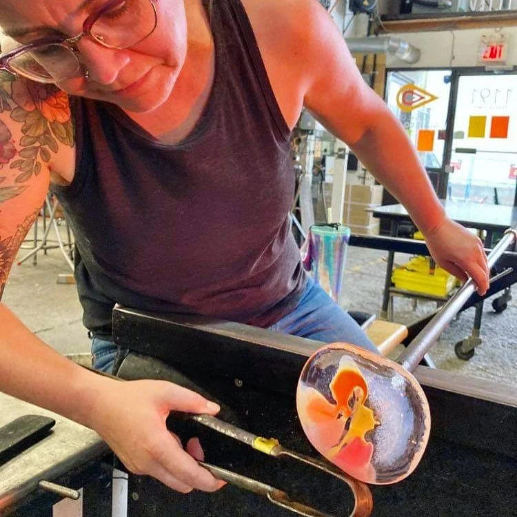 Sonya working with glass in a workshop. She is holding a heated piece of glass with tools, shaping a glass piece that has orange, yellow, and pink colors.