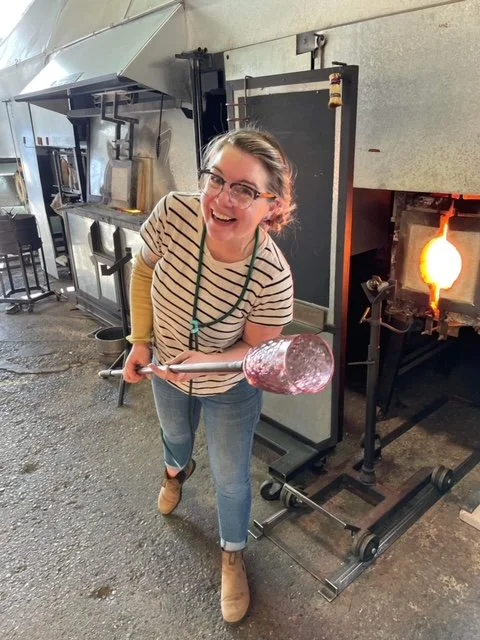 Sonya holding a glassblowing rod with a Gem cup, standing in a glassblowing studio with a furnace in the background.