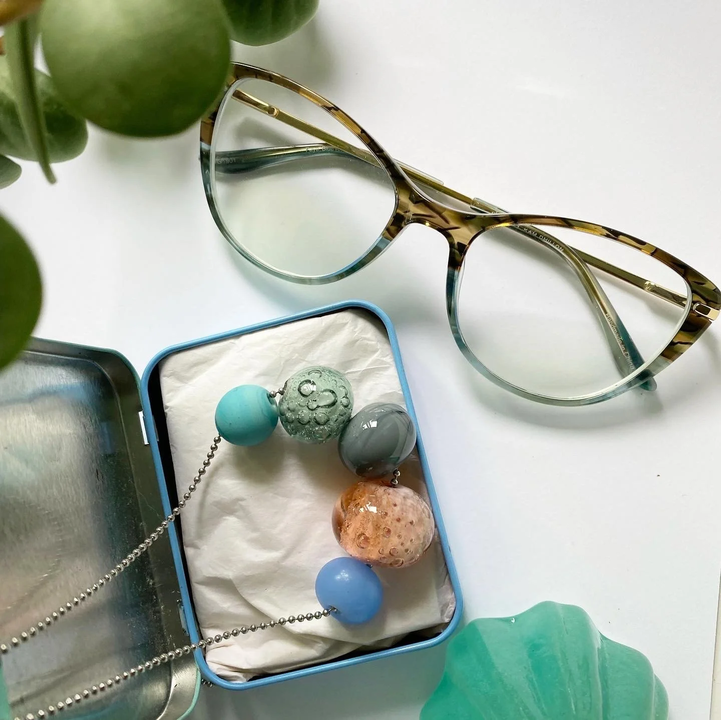 Pair of tortoise shell eyeglasses, colorful beaded necklace inside a blue jewelry box, and a green plant in the corner on a white surface.
