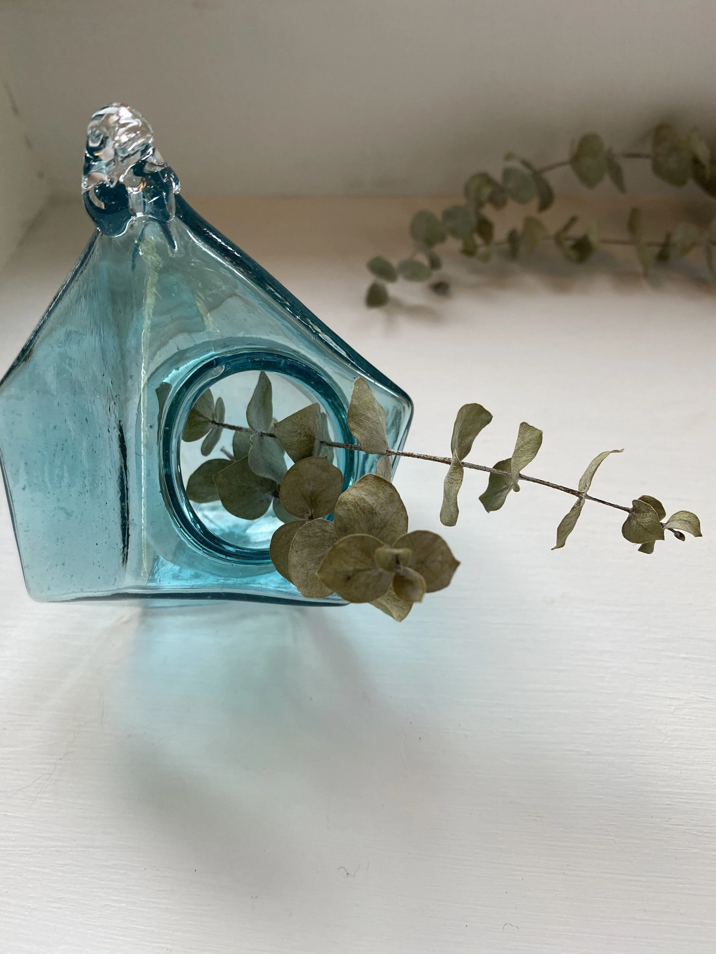 A turquoise glass geometric-shaped vase with a circular opening on its side, containing dried eucalyptus leaves, placed on a white surface.