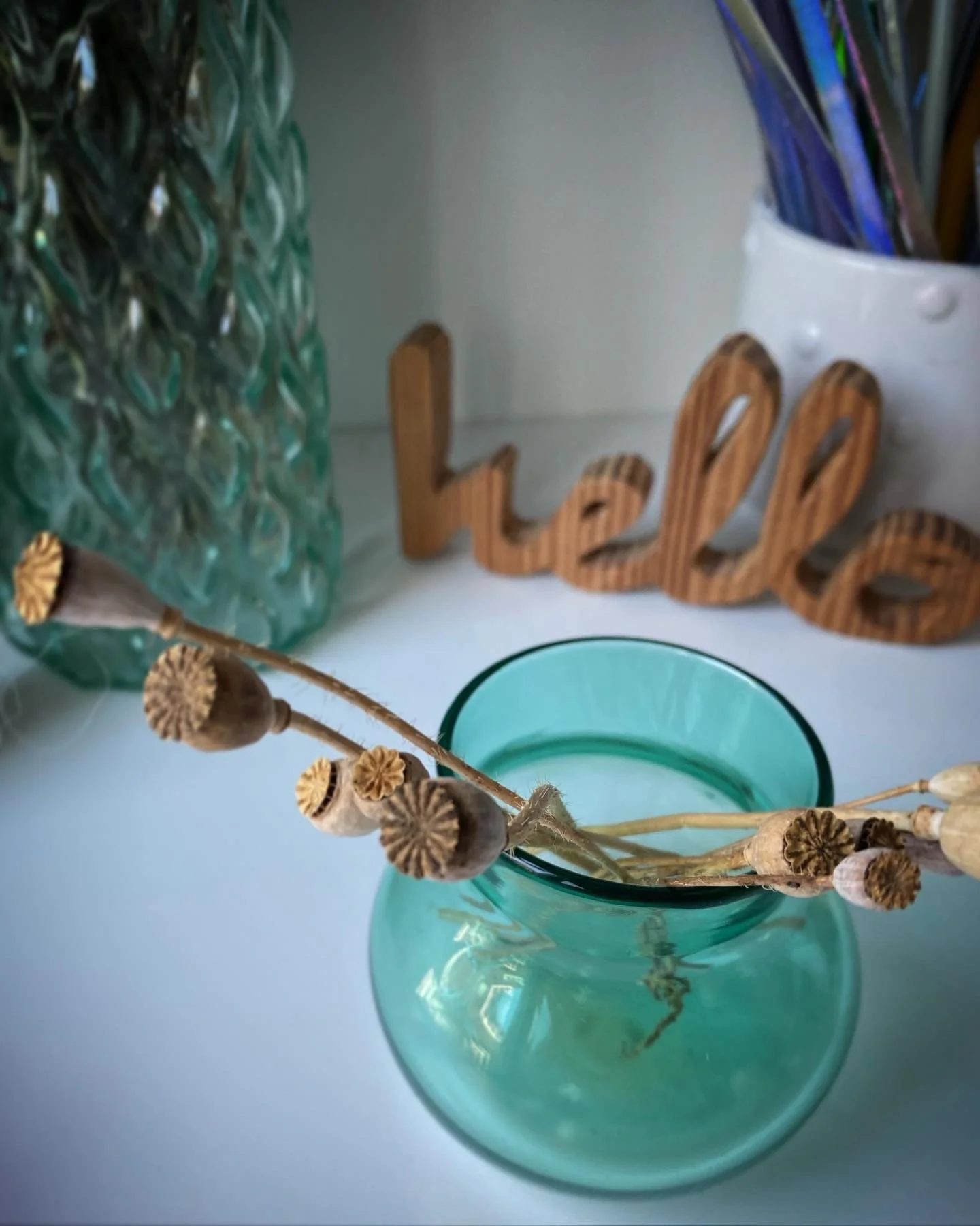A teal glass vase holding dried poppy seed pods and stems, positioned in front of a textured green glass container and a white container filled with colorful pens or markers on a white surface.