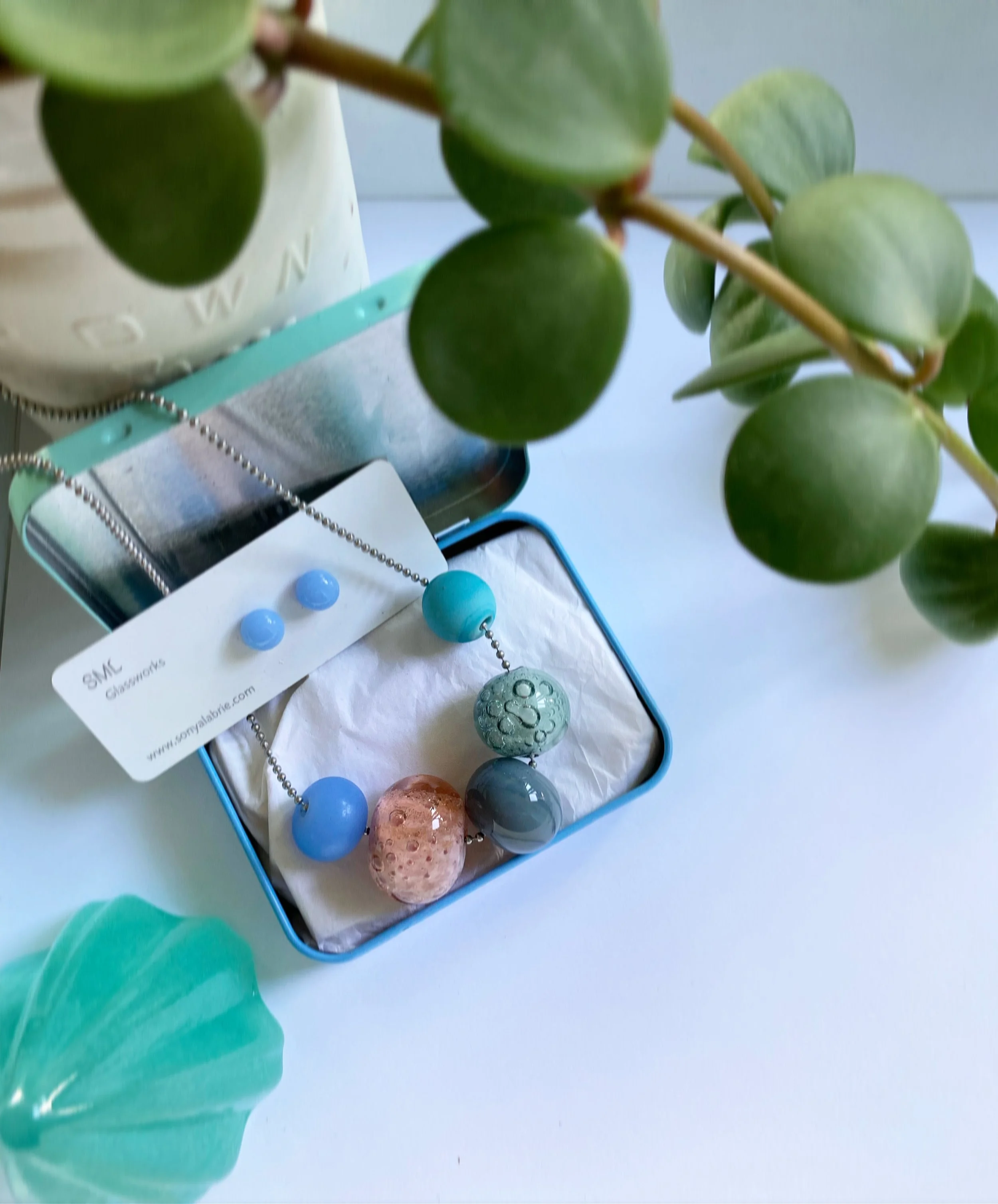 Close-up of a green leafy plant in a white pot with partially visible embossed letters, and a jewelry display tray with blue, green, and pink beads and a necklace on a white surface.