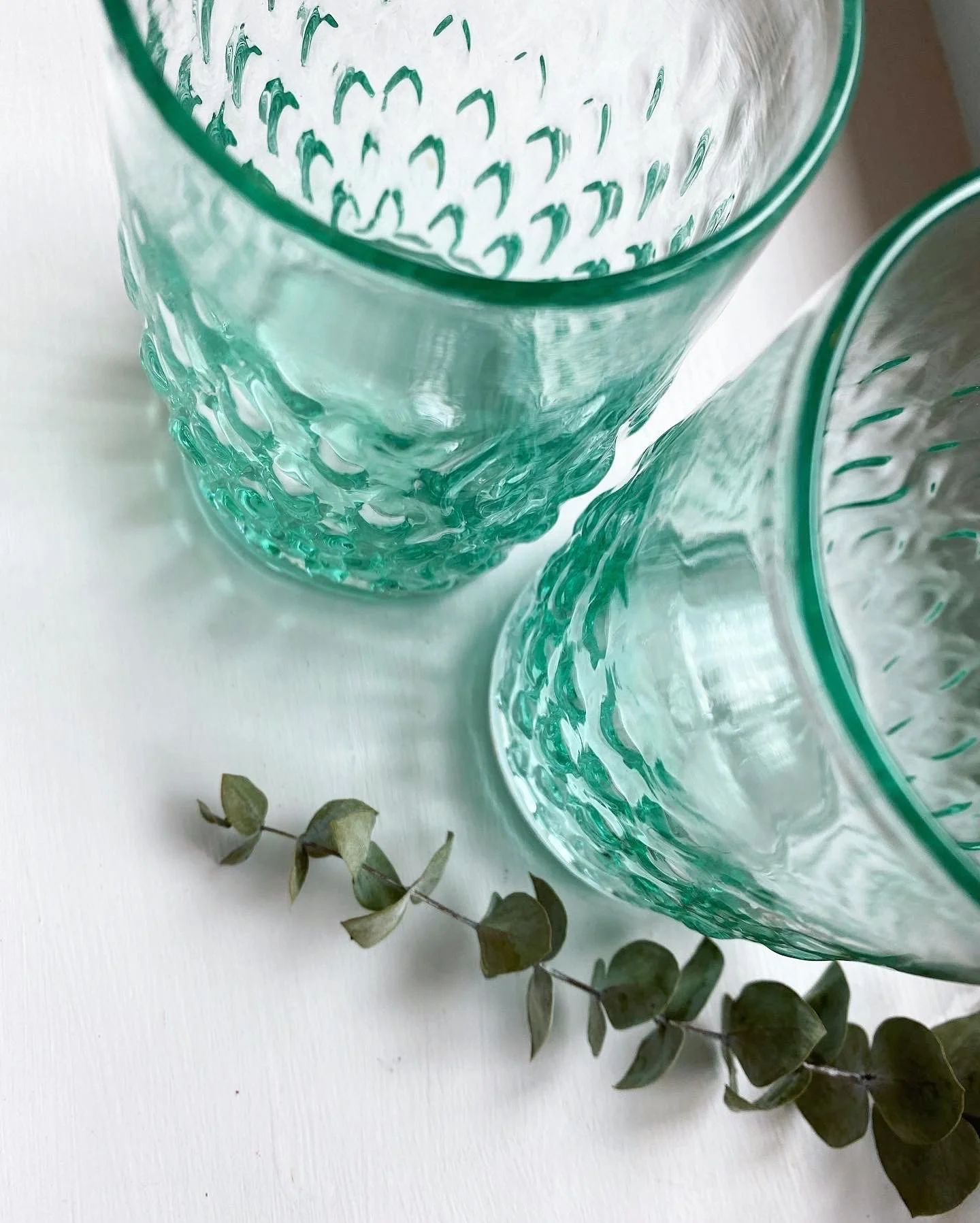 Two textured gem cup glass with a green tint and a eucalyptus branch on a white surface.