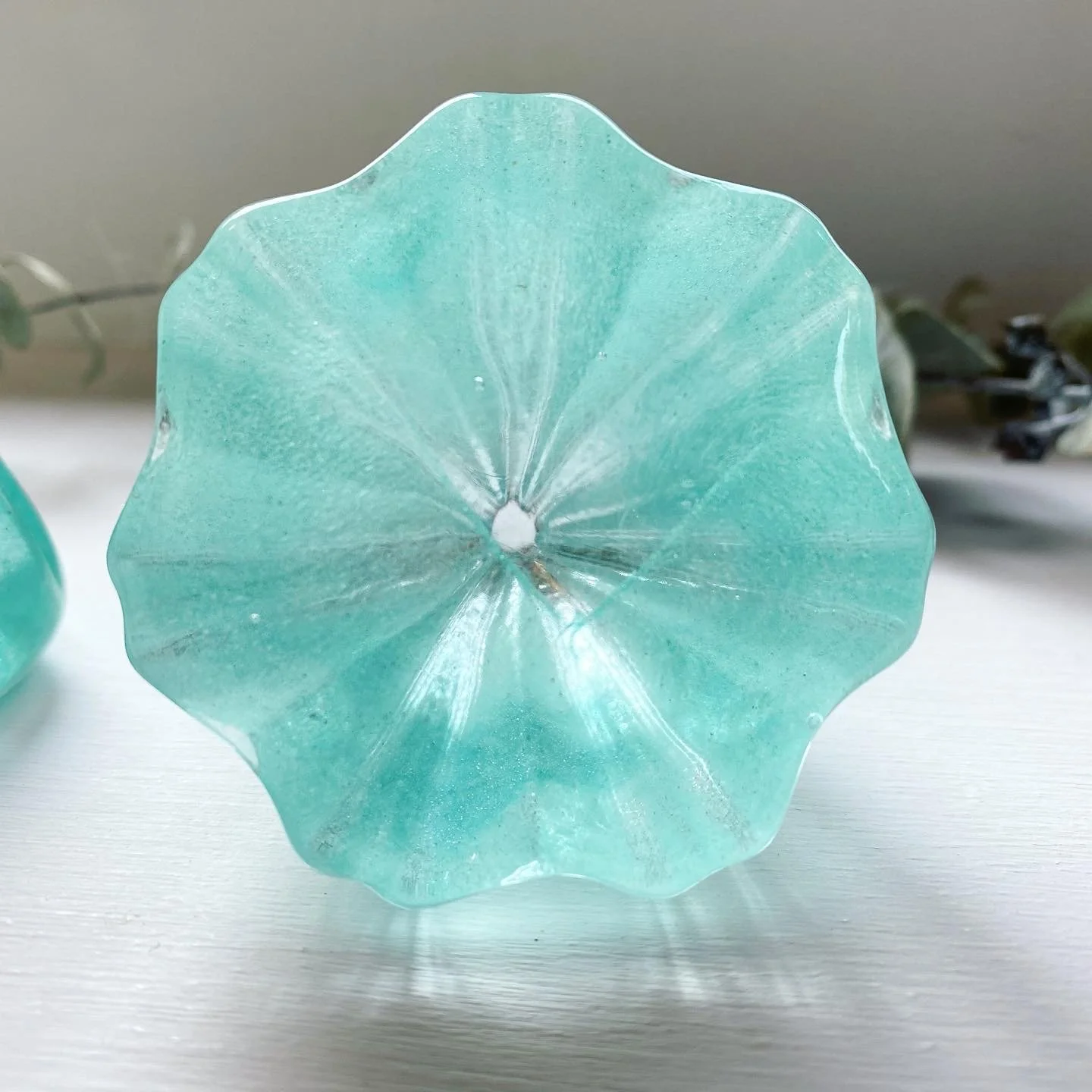 Close-up of a light turquoise glass vase with a wavy, flower petal-like edge, placed on a white surface with a blurred background, including some green and dark-colored plants.