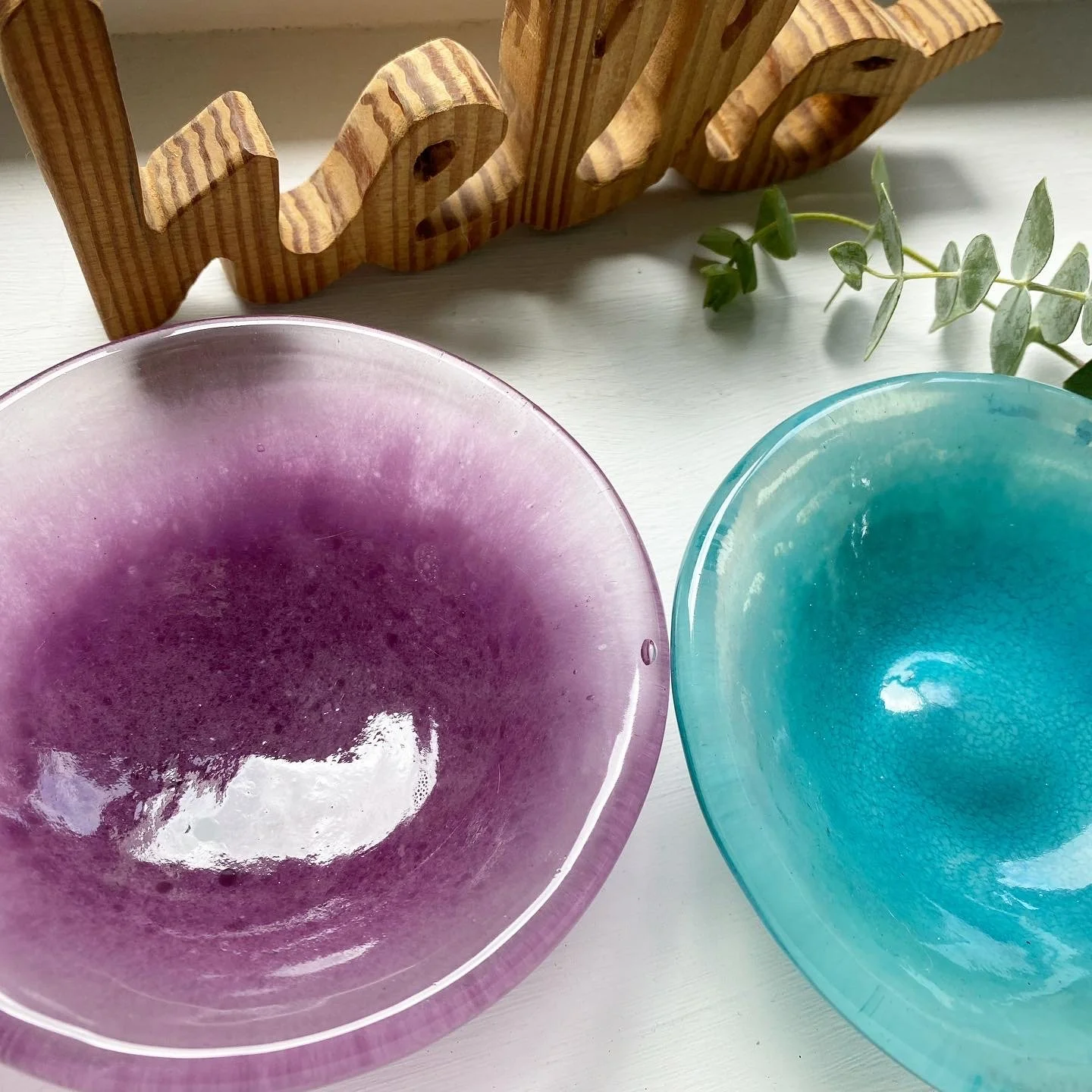 Close-up of two colorful glass bowls, one pink and one teal, with bubble textures, placed on a white surface. A wooden word decoration spelling "love" and a green leafy plant are in the background.