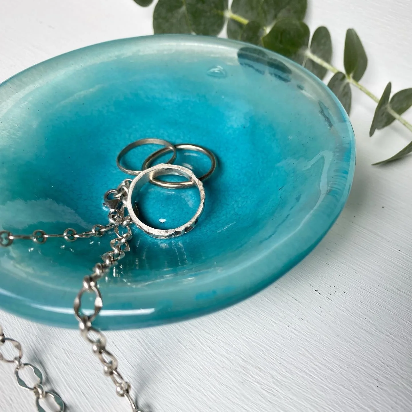A blue glass dish with a silver necklace and rings inside, on a white surface with green leaves in the background.