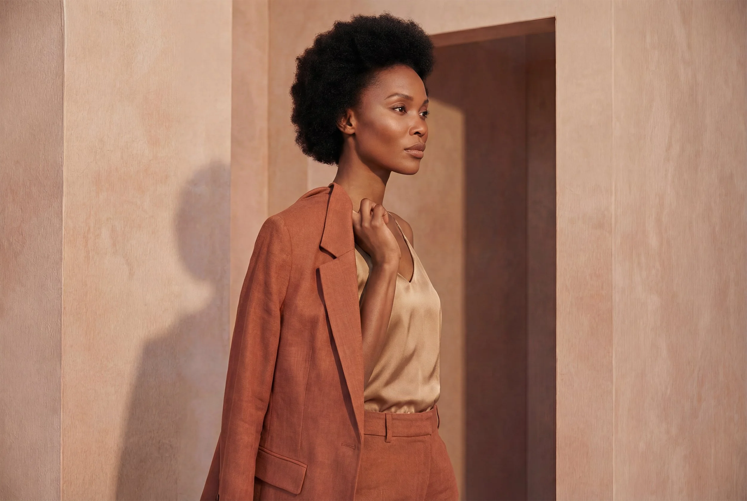 Donna afroamericana con abito beige e giacca marrone in uno spazio dai colori caldi.