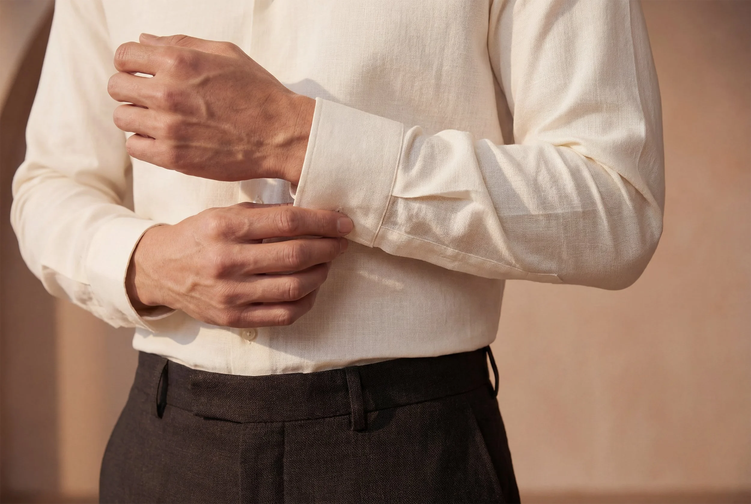 Una persona con camicia chiara che si sistema il polsino della manica, indossa pantaloni grigi, fondo neutro.