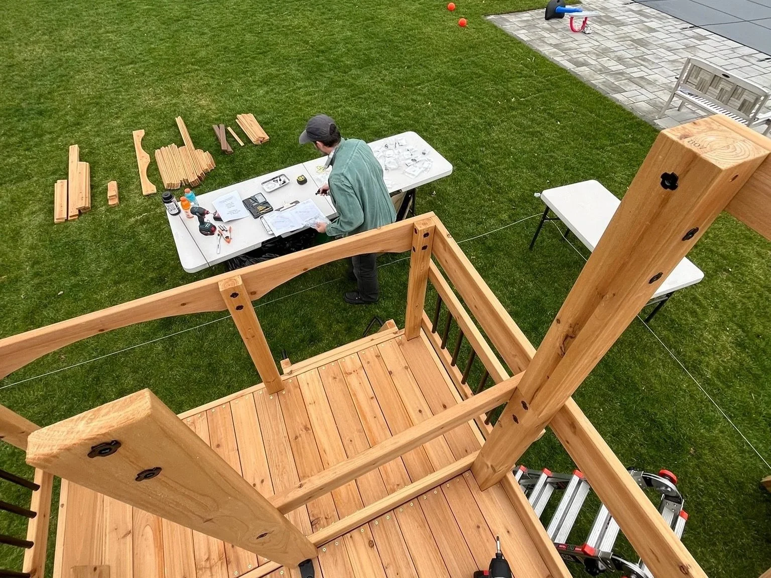 Person working on assembling a wooden outdoor playset or treehouse on a grassy lawn with tools and wood pieces on a table.