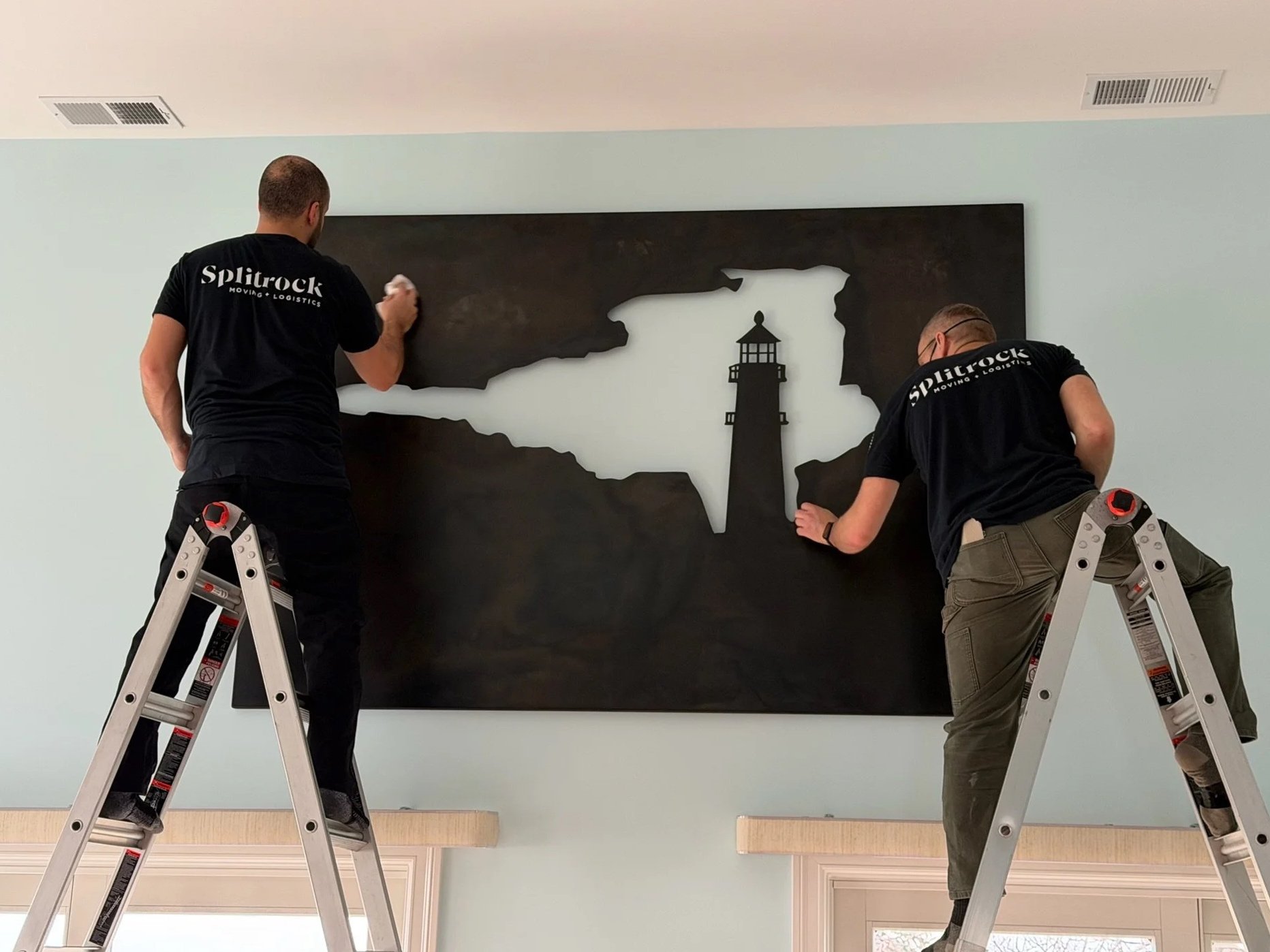 Two workers on ladders installing a large wall art piece featuring a cutout silhouette of the state of Michigan with a lighthouse inside the silhouette