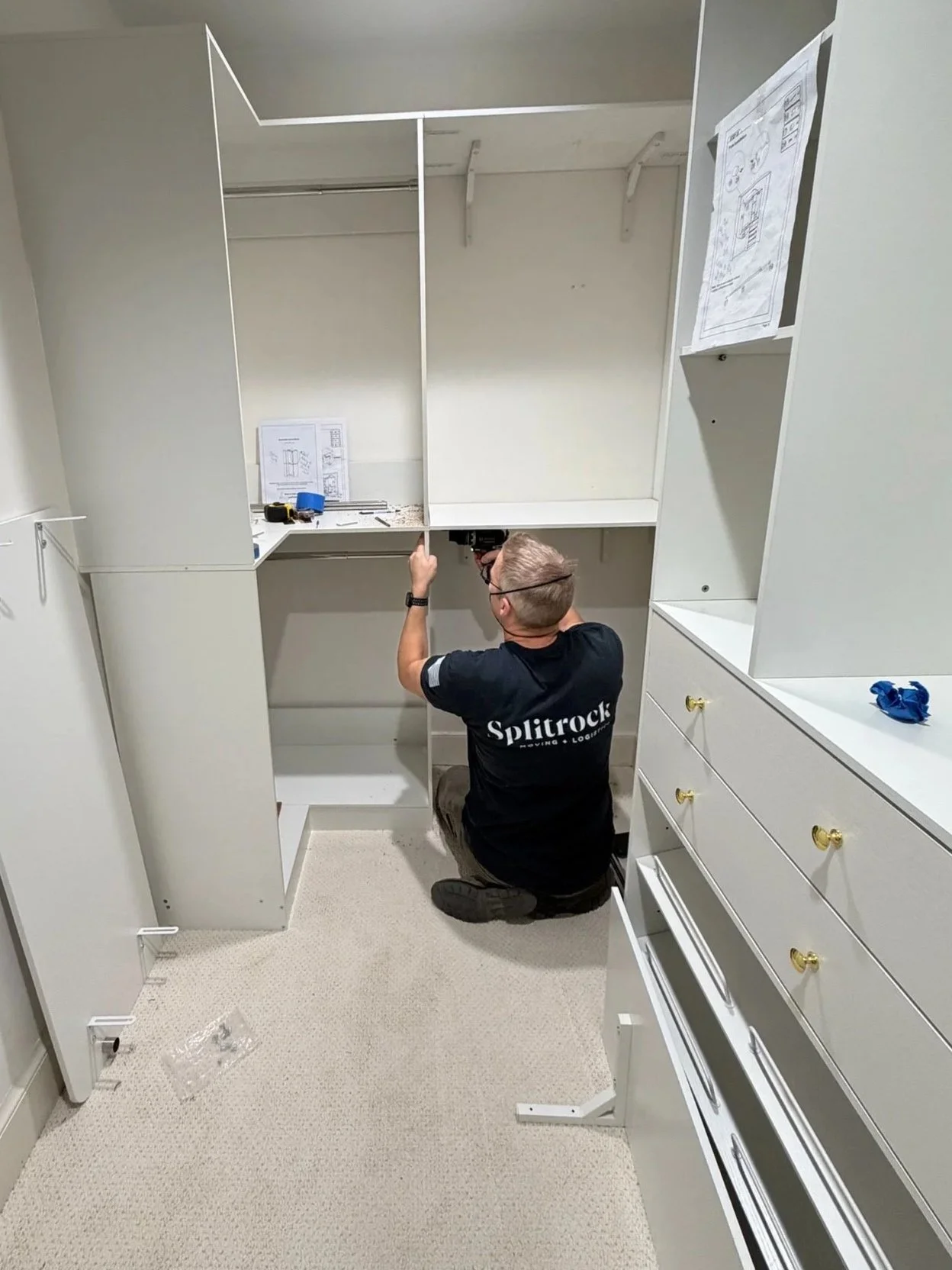 A person kneeling on the floor installing and assembling a furniture system, with tools and instructions visible on the shelves.