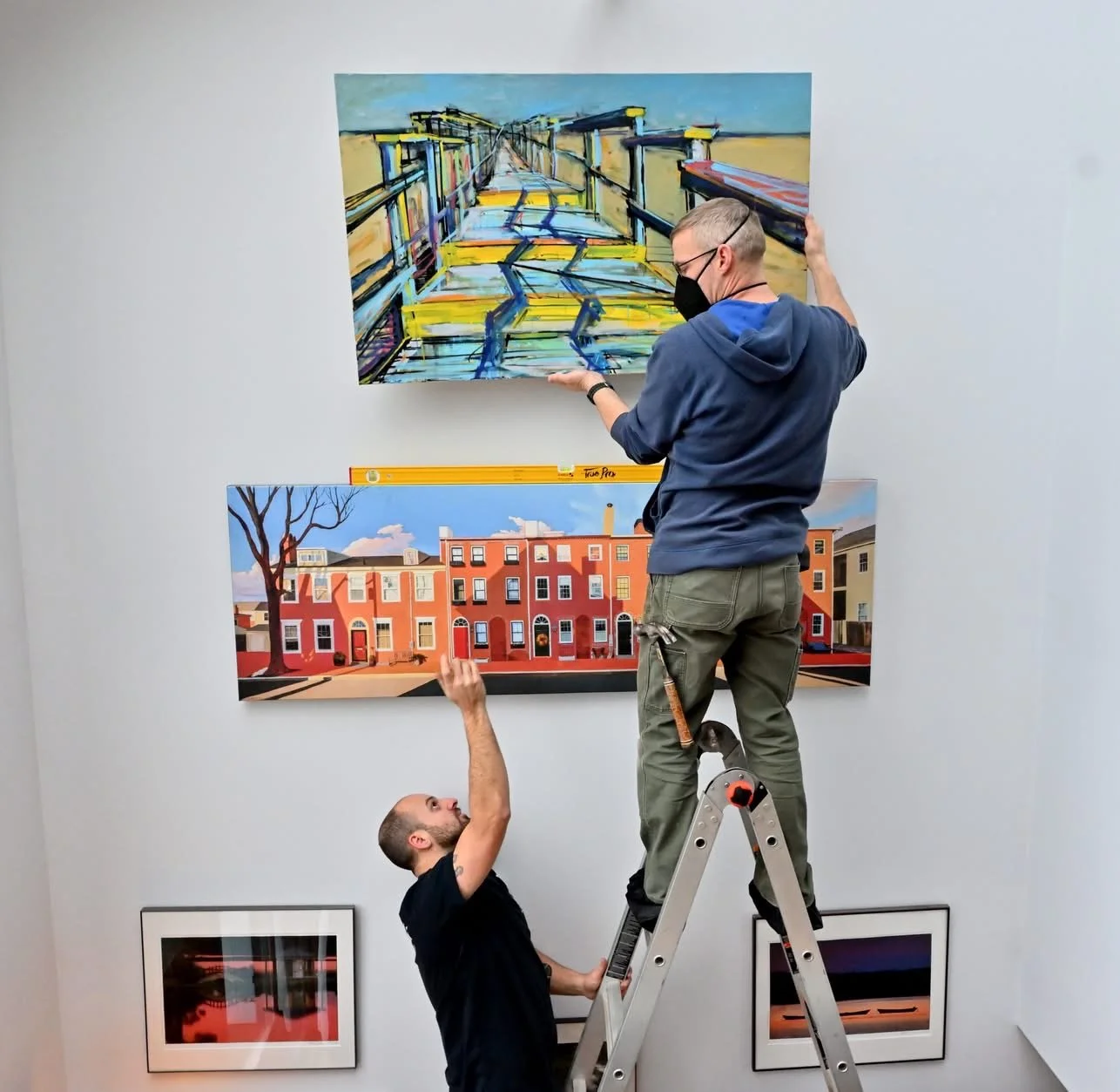Two men installing and adjusting artwork on a gallery wall, one standing on a ladder and one supporting from below.