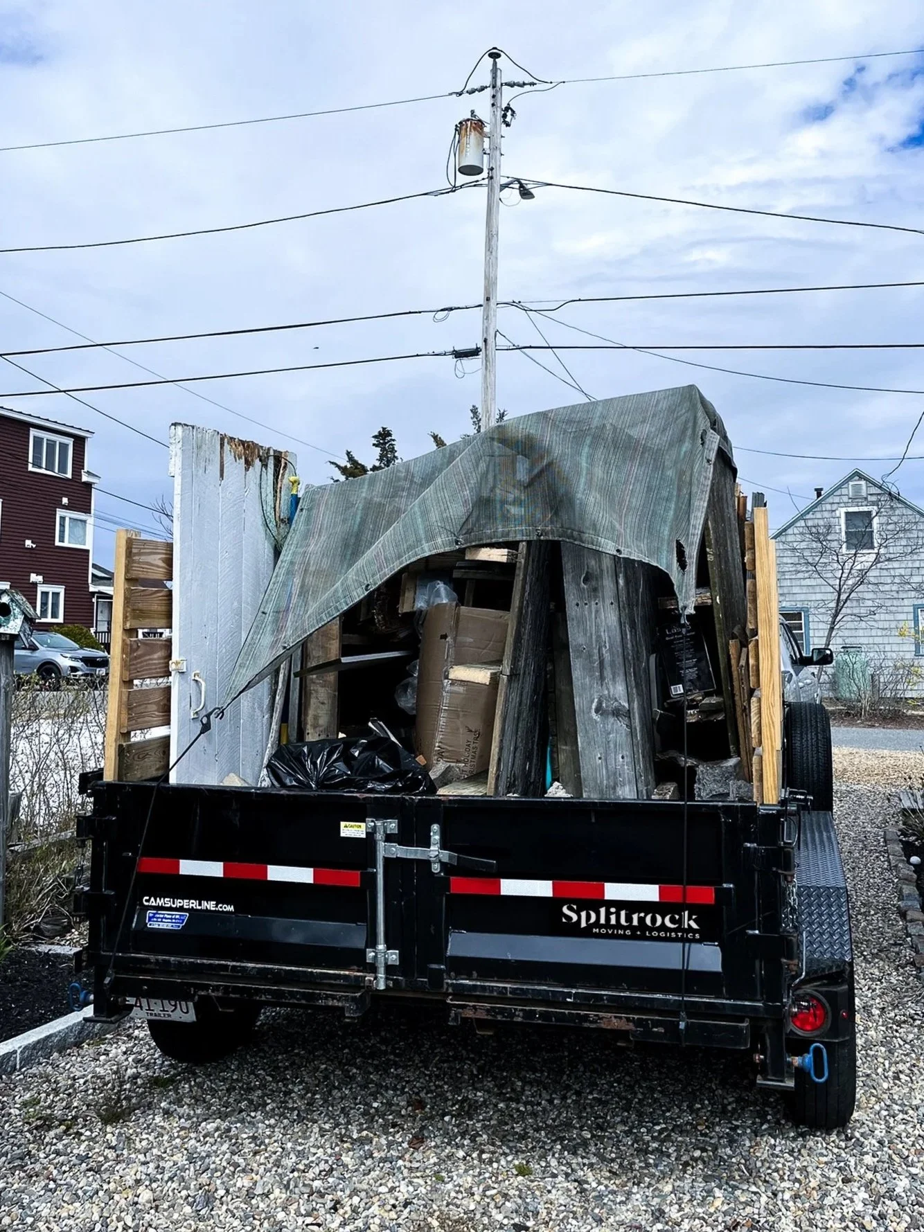 Trailer loaded with construction debris, covered partially with a green tarp, parked on gravel near residential houses.
