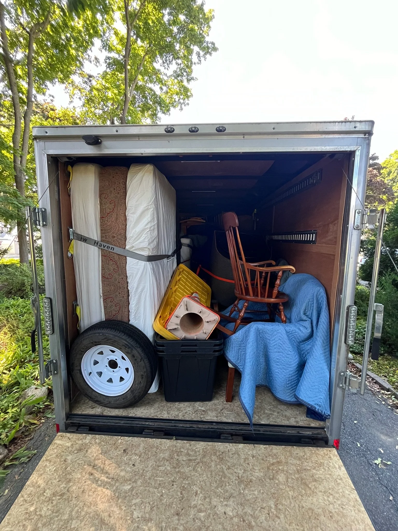 The inside of an enclosed moving truck filled with furniture and household items, including a mattress, chairs, a tire, a plastic bin, and a blue quilted blanket.