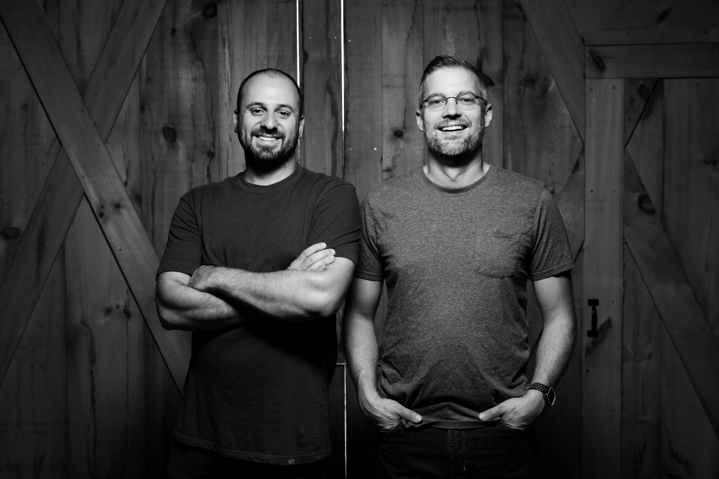 Two smiling men with beards standing side by side in front of a wooden wall. The man on the left is crossing his arms, and the man on the right has his hands in his pockets.