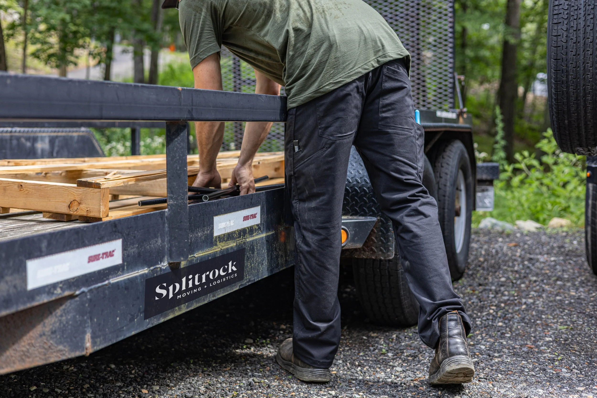 Man loading wood and equipment onto a black trailer in a wooded area.