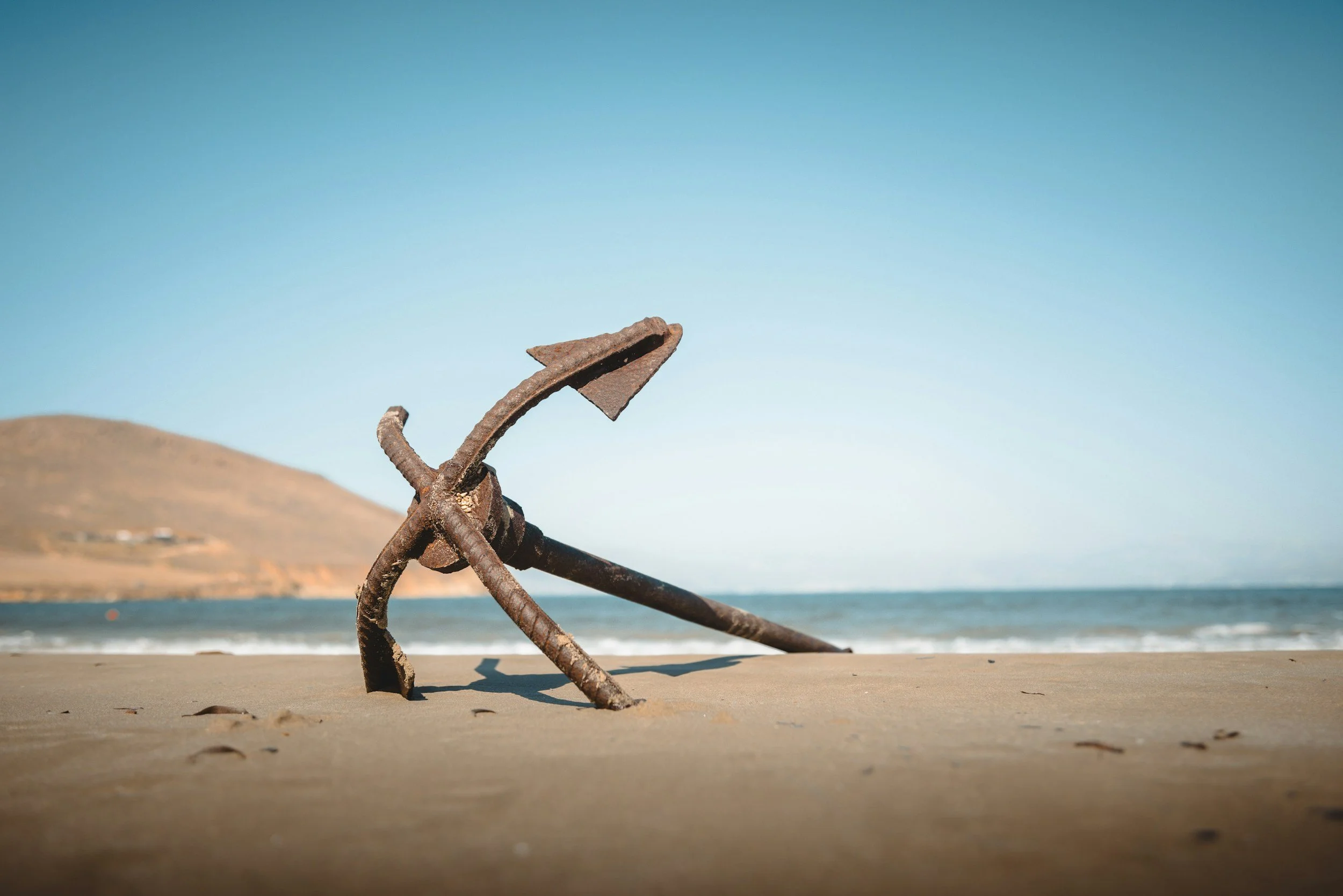 Old rusty anchor on sandy beach with ocean and hills in the background under clear blue sky.
