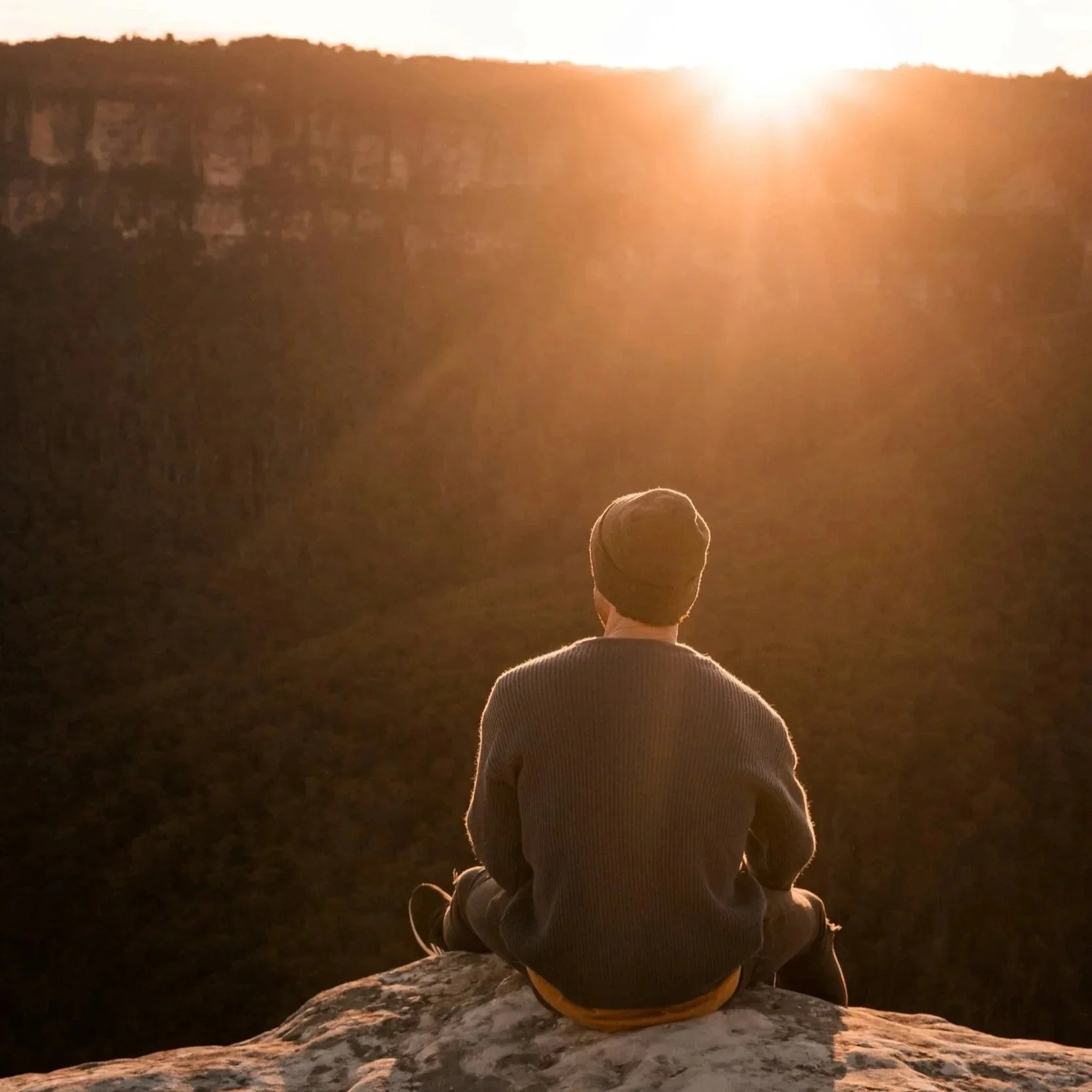A person sitting cross-legged on a rock, overlooking a deep canyon during sunset, with the sun low on the horizon casting a warm glow.