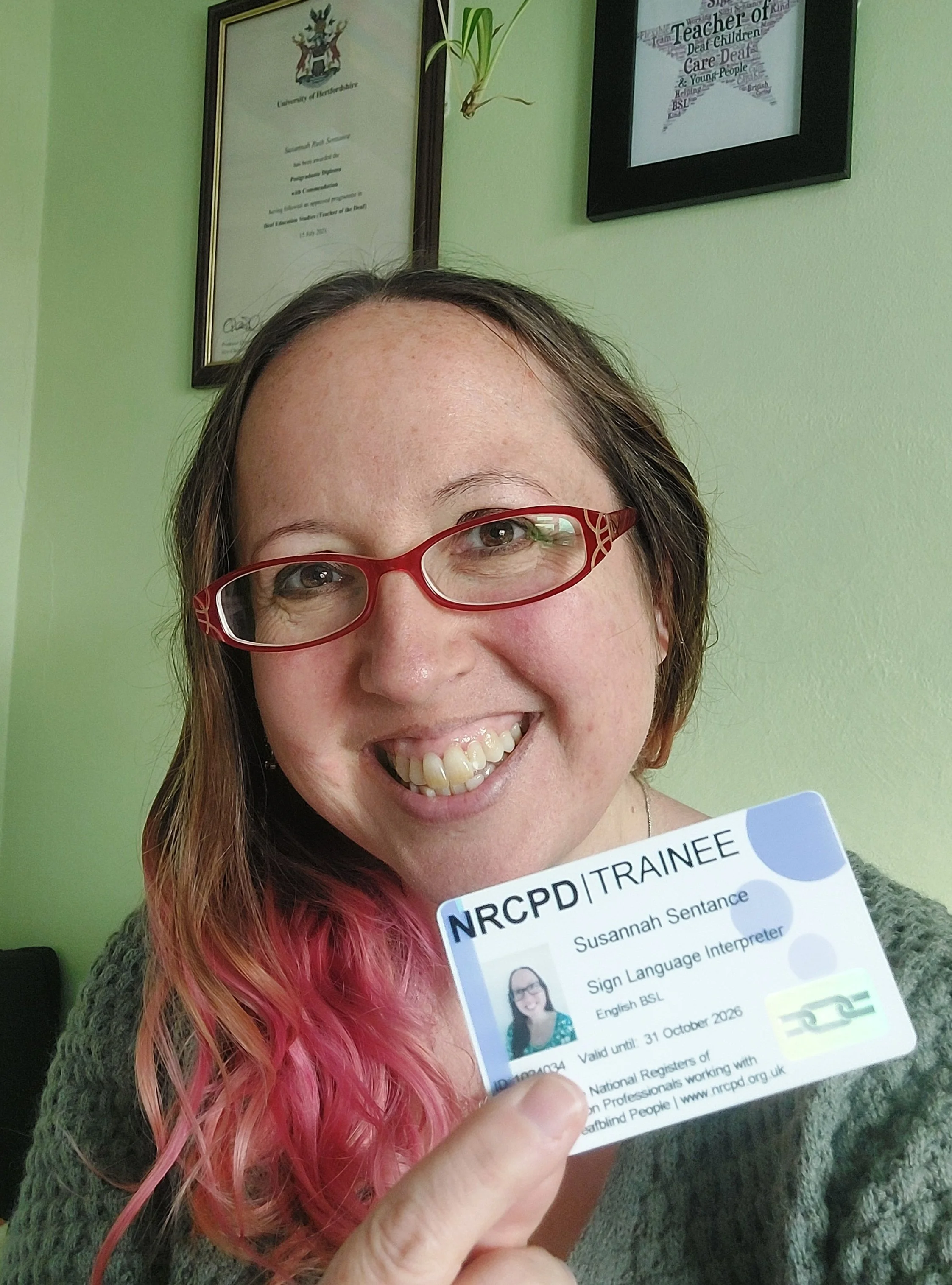 A smiling Suzi with red glasses and a green dress sits in front of a pale green wall, holding a badge that says NRCPD Trainee.