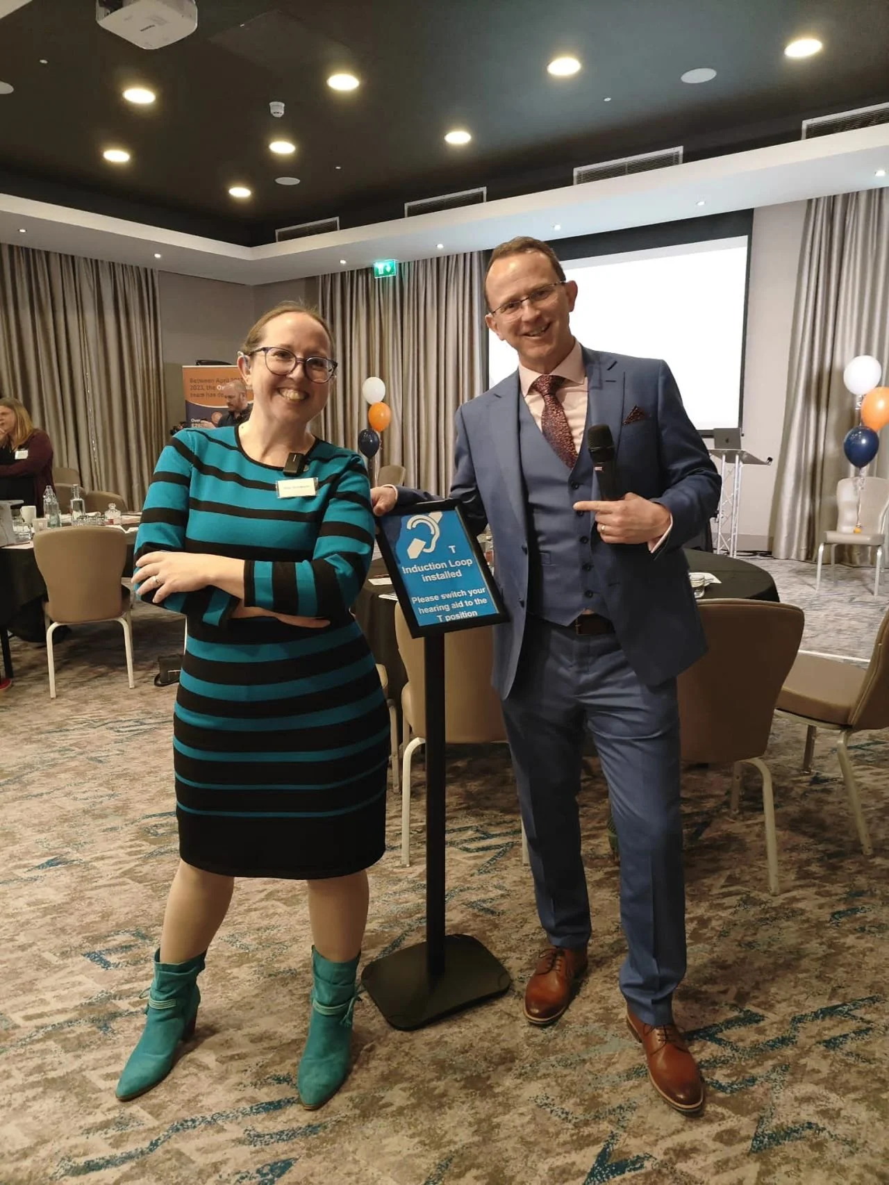 Suzi, in a teal and black striped dress, stands in a conference room with a man in a suit holding a microphone. In between them is a sign saying induction loop installed.
