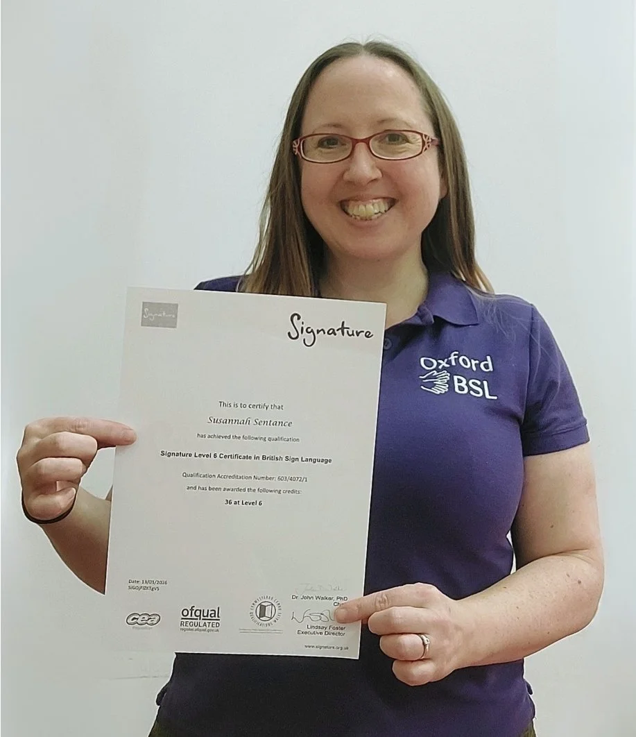 Suzi stands against a white wall and is holding an A4 certificate that says Signature Level 6 Certificate in British Sign Language. She is wearing red glasses, a purple polo shirt with the Oxford BSL logo and a big smile!