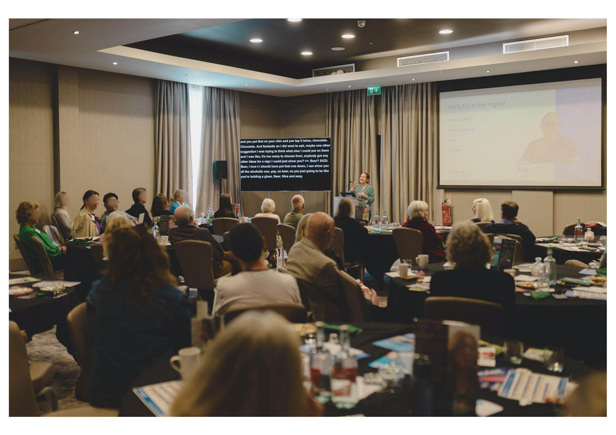 A conference room with lots of people sitting at circular tables with drinks and leaflets scattered. Suzi stands in between a captioning display and a projector screen demonstrating a BSL sign.