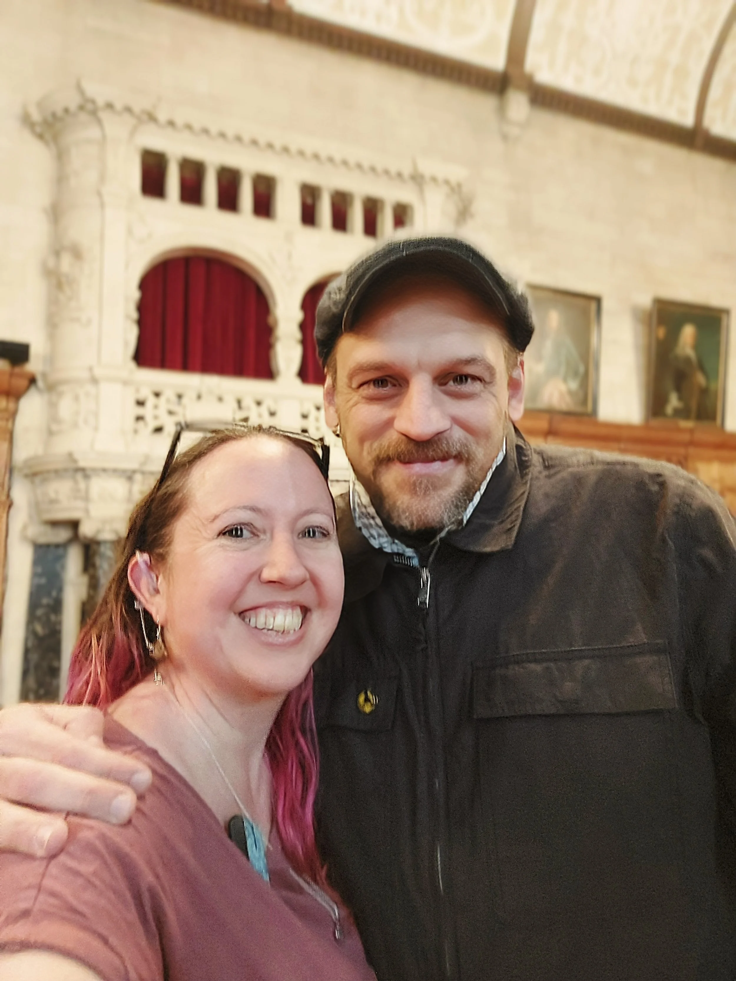 Suzi, wearing a purple t-shirt, stands with Matthew Gurney, wearing a flat cap and jacket. They are both smiling and looking at the camera.