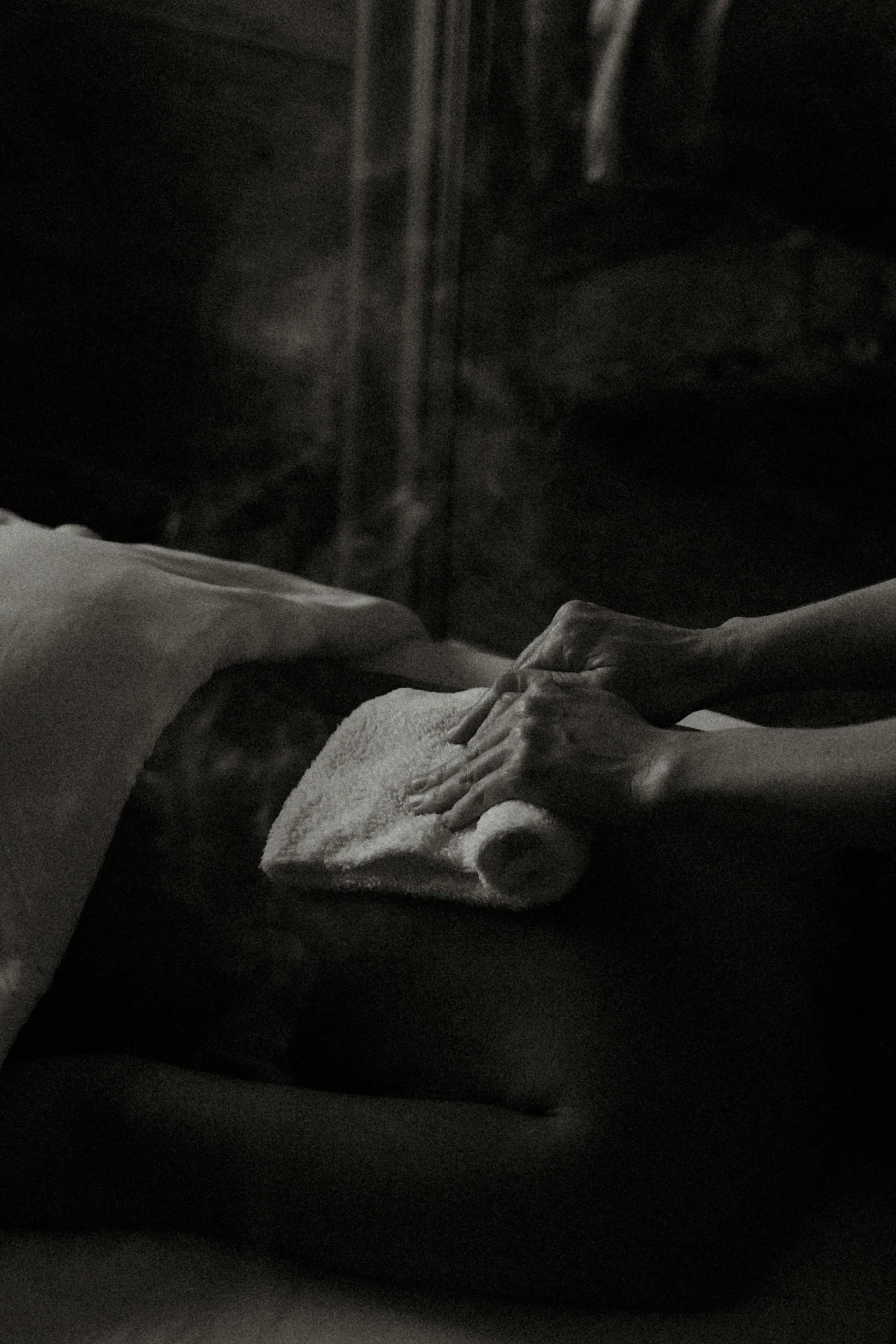 Person receiving a massage on their back in a dark room, with hands pressing down on their back, a towel rolled up beside them, and a pillow under their head.