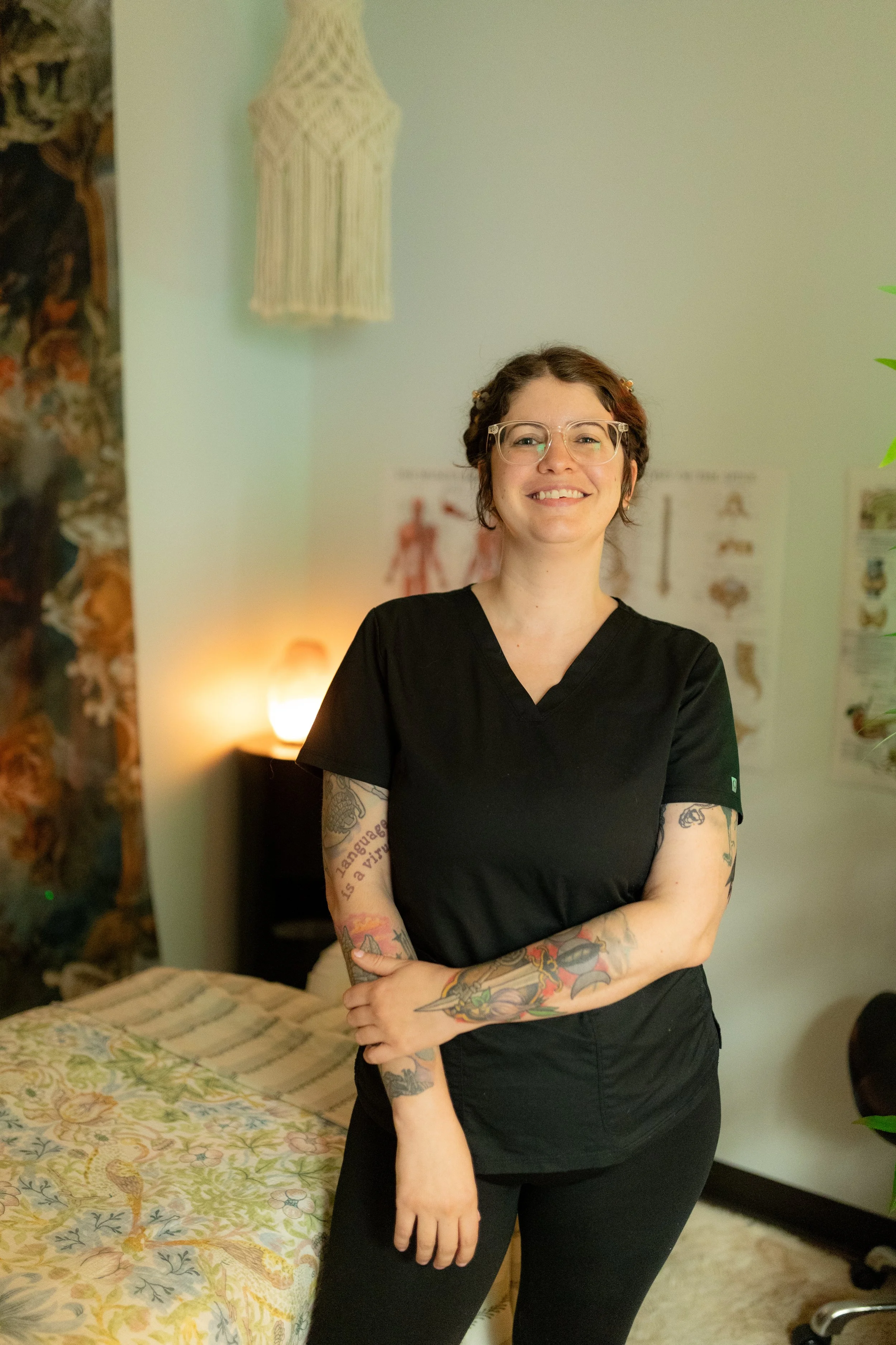 A smiling woman with glasses, tattoos on her arms, wearing a black scrubs top, standing in a room with a bed, posters, and a lamp.